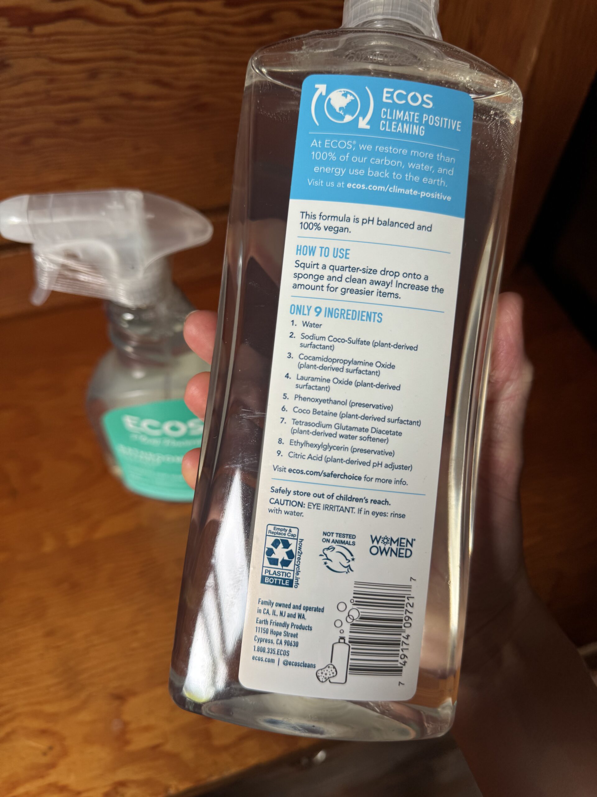 Hand holding a clear bottle of ECOS cleaner showing the back label with product information, ingredients list, usage instructions, and a spray bottle in the background.