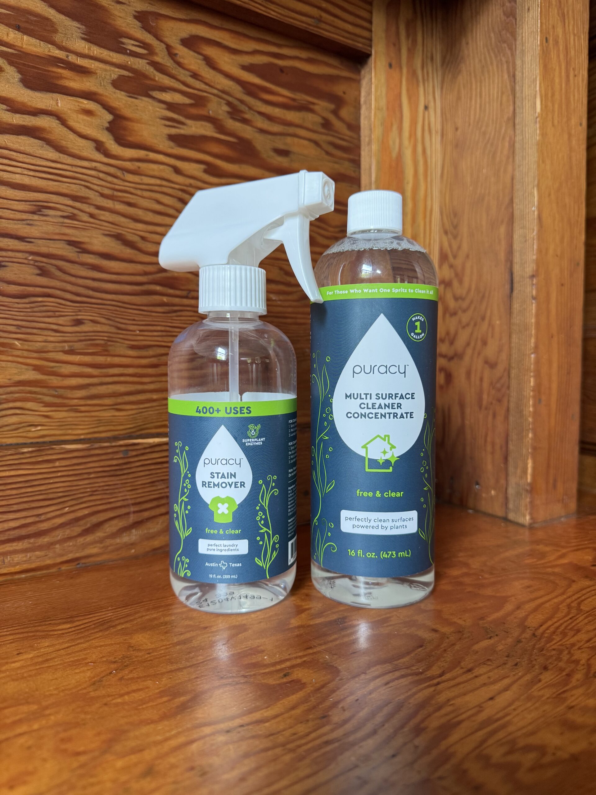 Two bottles of Puracy cleaning products, one is a stain remover with a spray nozzle, and the other is a multi-surface cleaner concentrate, both on a wooden surface.