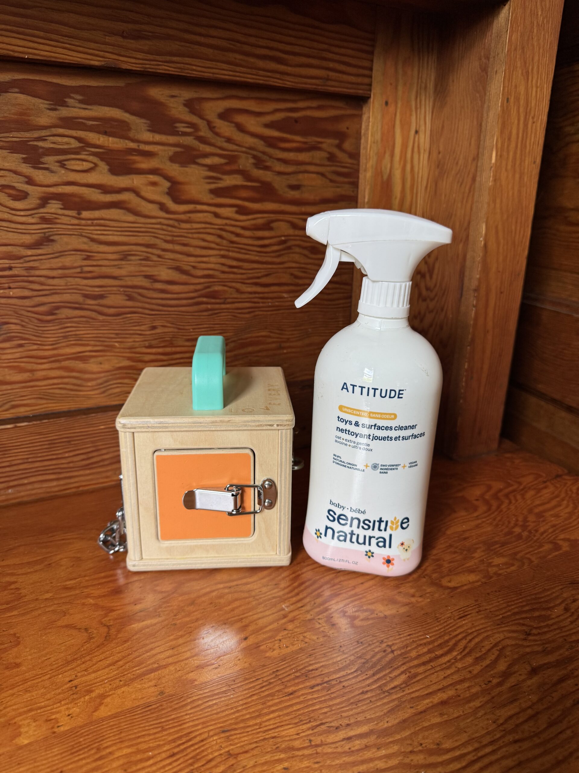 A wooden lock box with a turquoise handle sits next to a bottle of Attitude toy and surface cleaner on a wooden shelf.