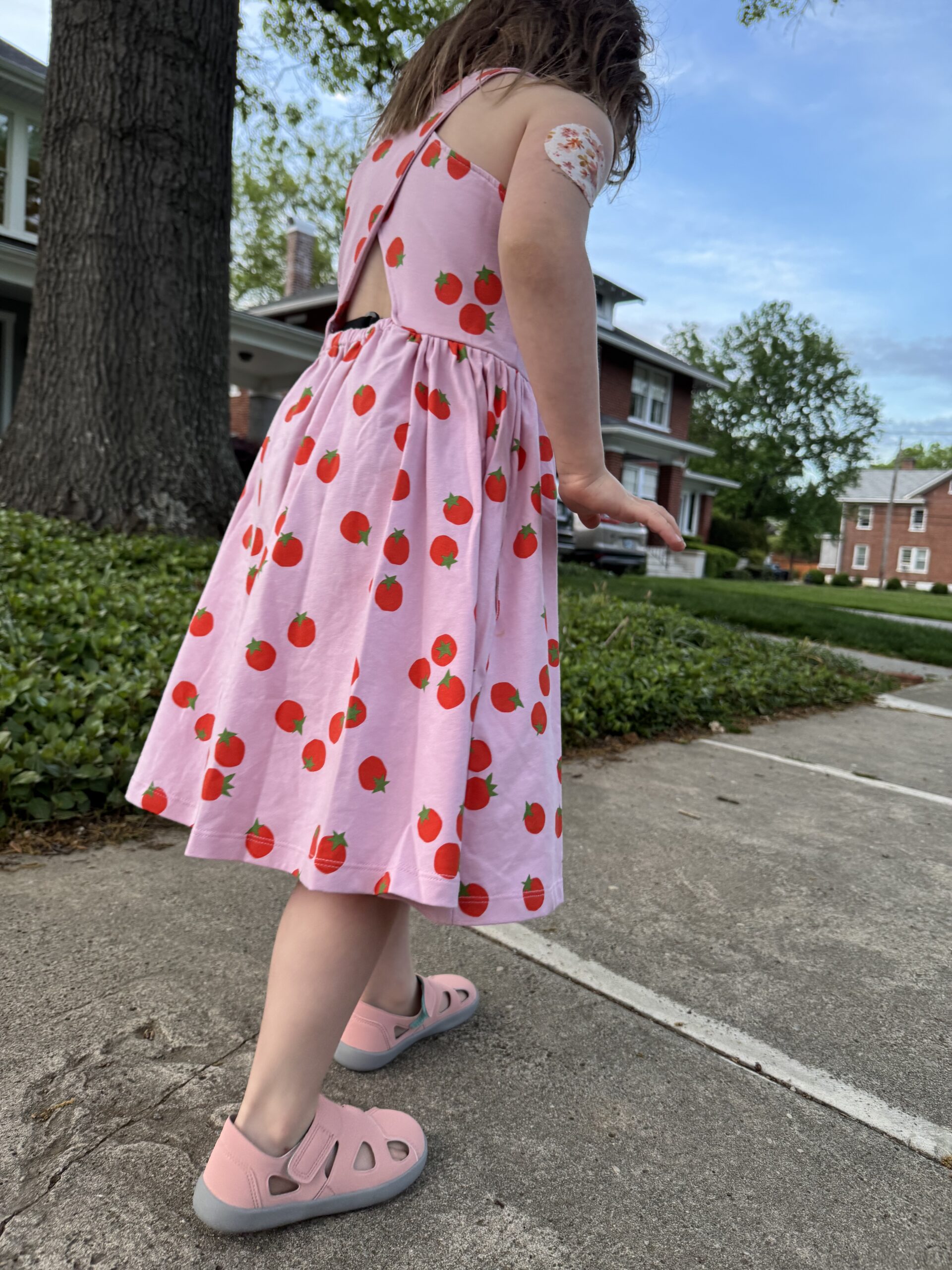 A child in a pink dress with a red fruit pattern and pink sandals walks on a sidewalk, with a bandage on their upper arm. Trees and houses are visible in the background.