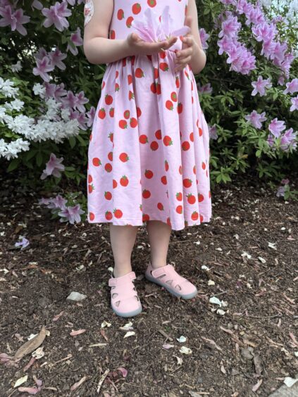 A young child in a pink dress with a strawberry print stands on mulch in front of blooming azalea bushes, with only the lower half of the body visible.