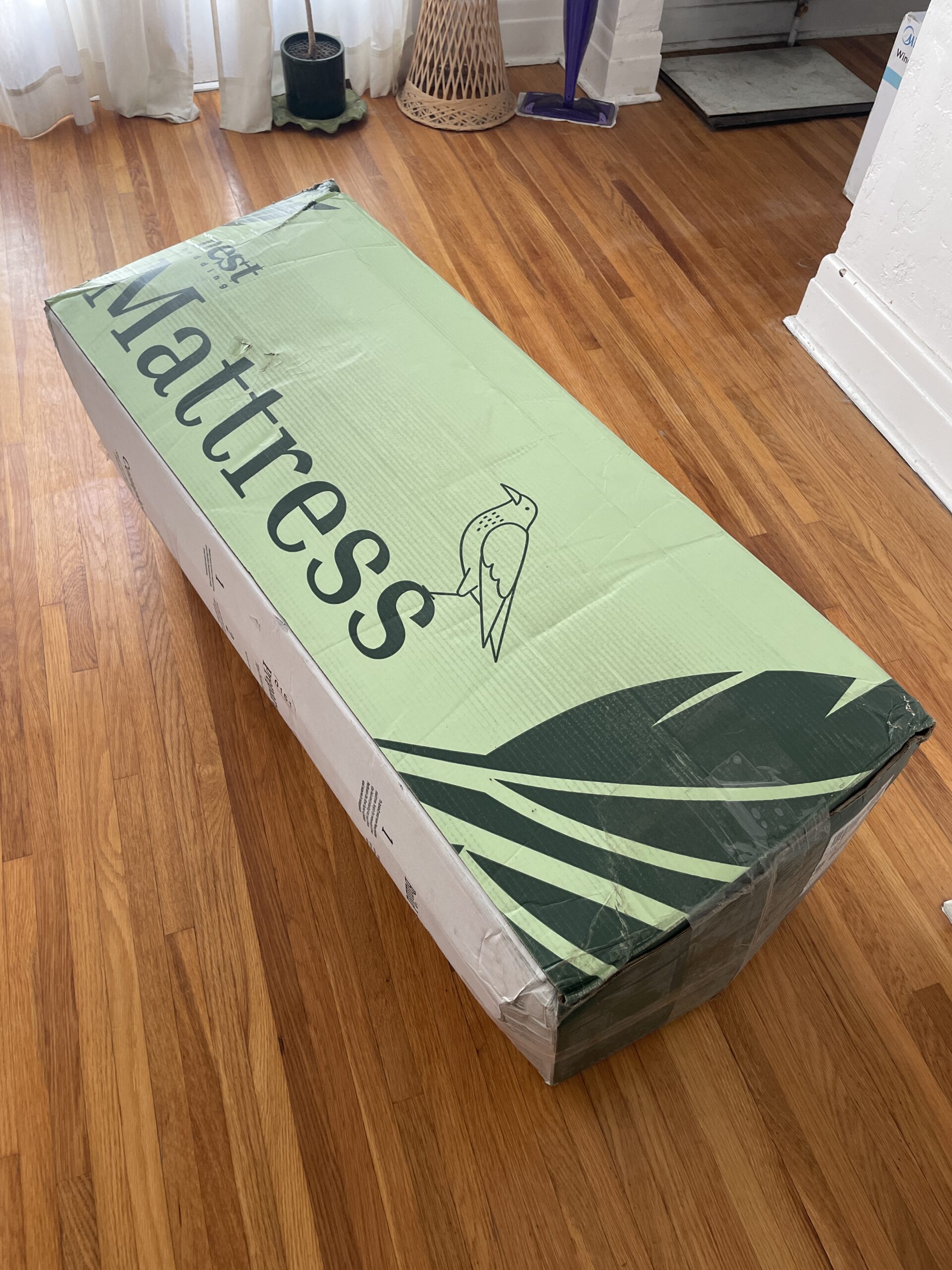 A green and white box labeled "mattress" is standing on a wooden floor. The box features a bird illustration.