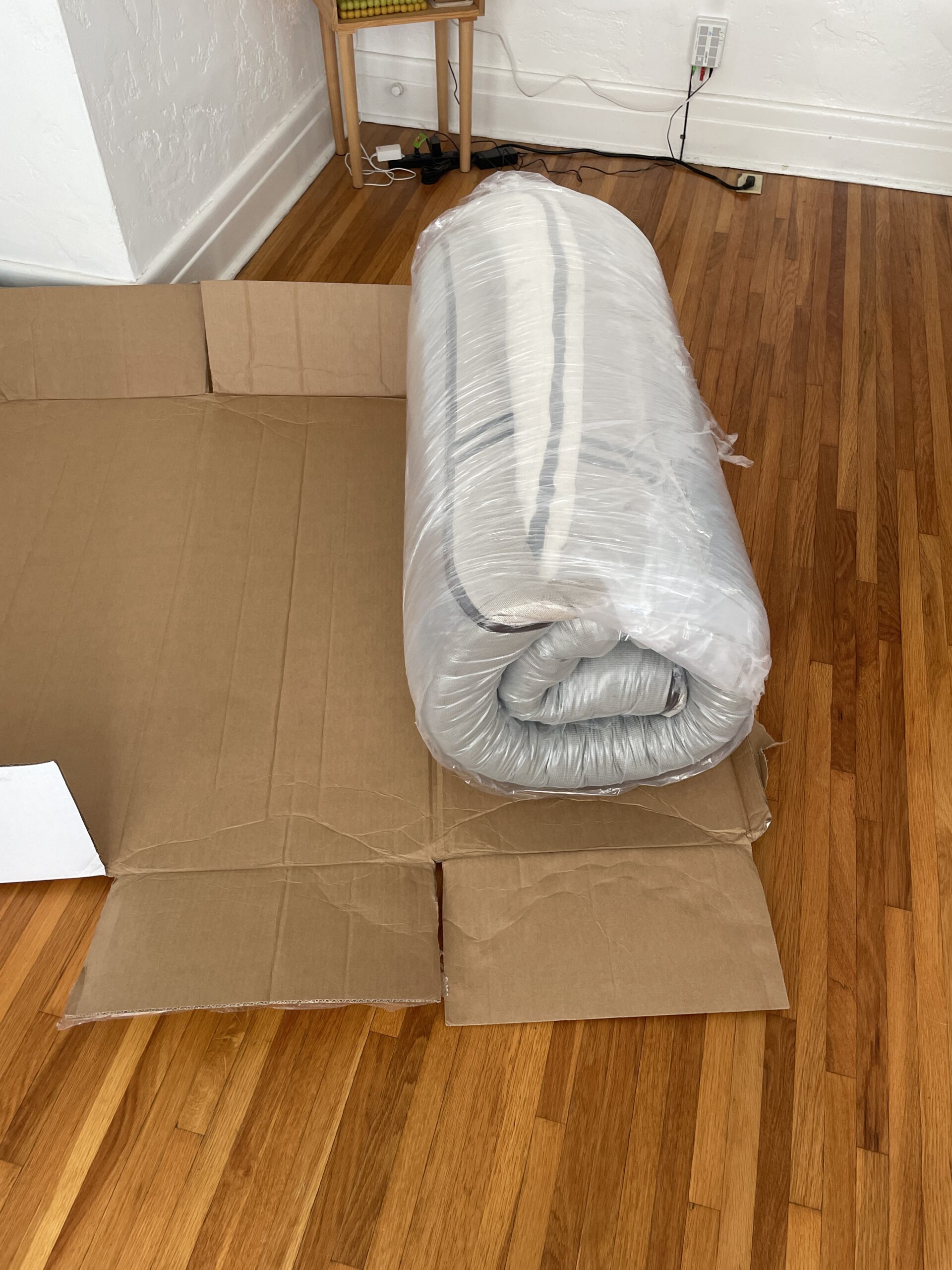 A rolled-up mattress wrapped in plastic lies on an open cardboard box on a wooden floor.