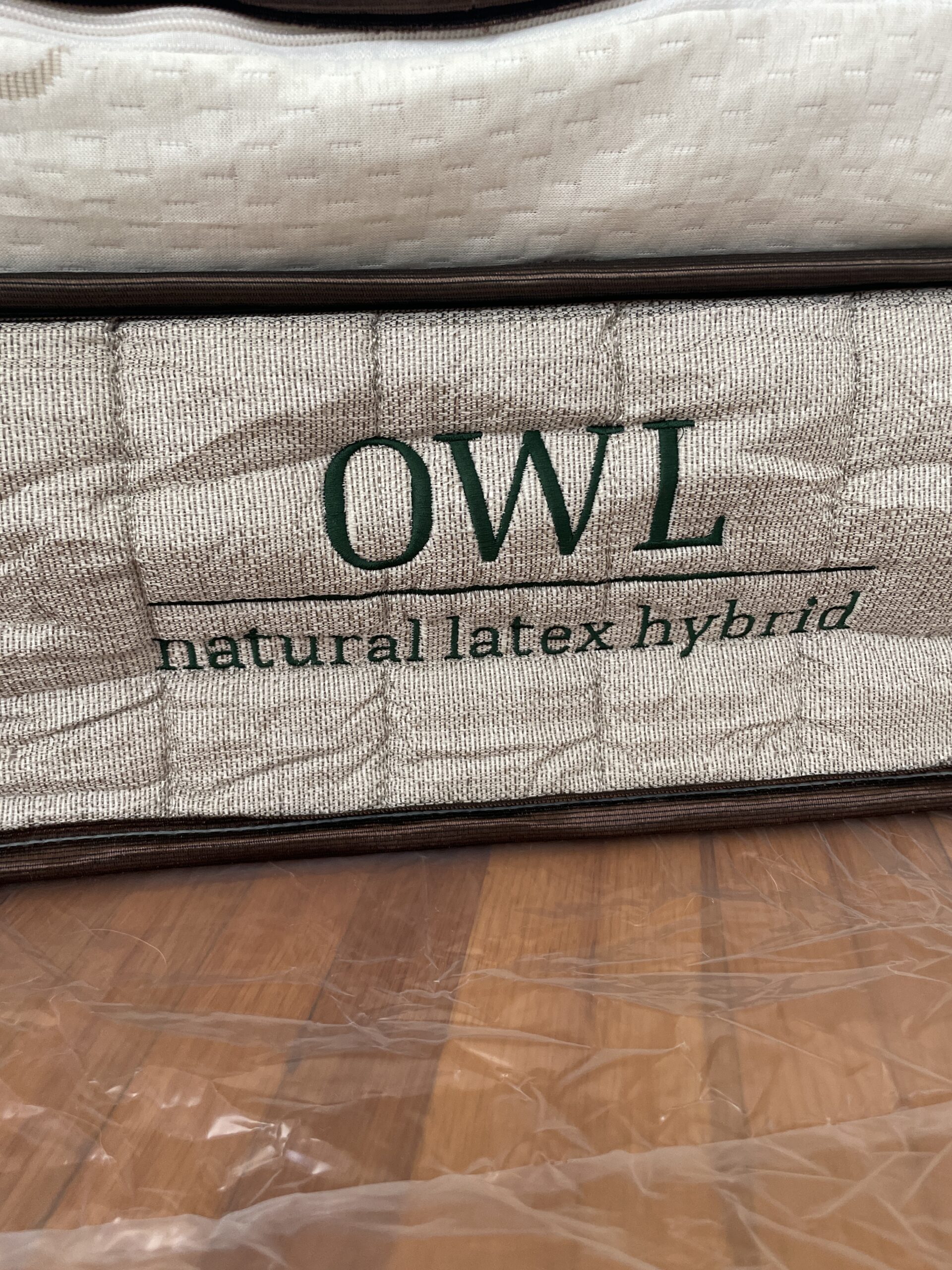 Close-up of a mattress label showing "OWL natural latex hybrid" in green text on a textured fabric background. The mattress is placed on a wooden floor.