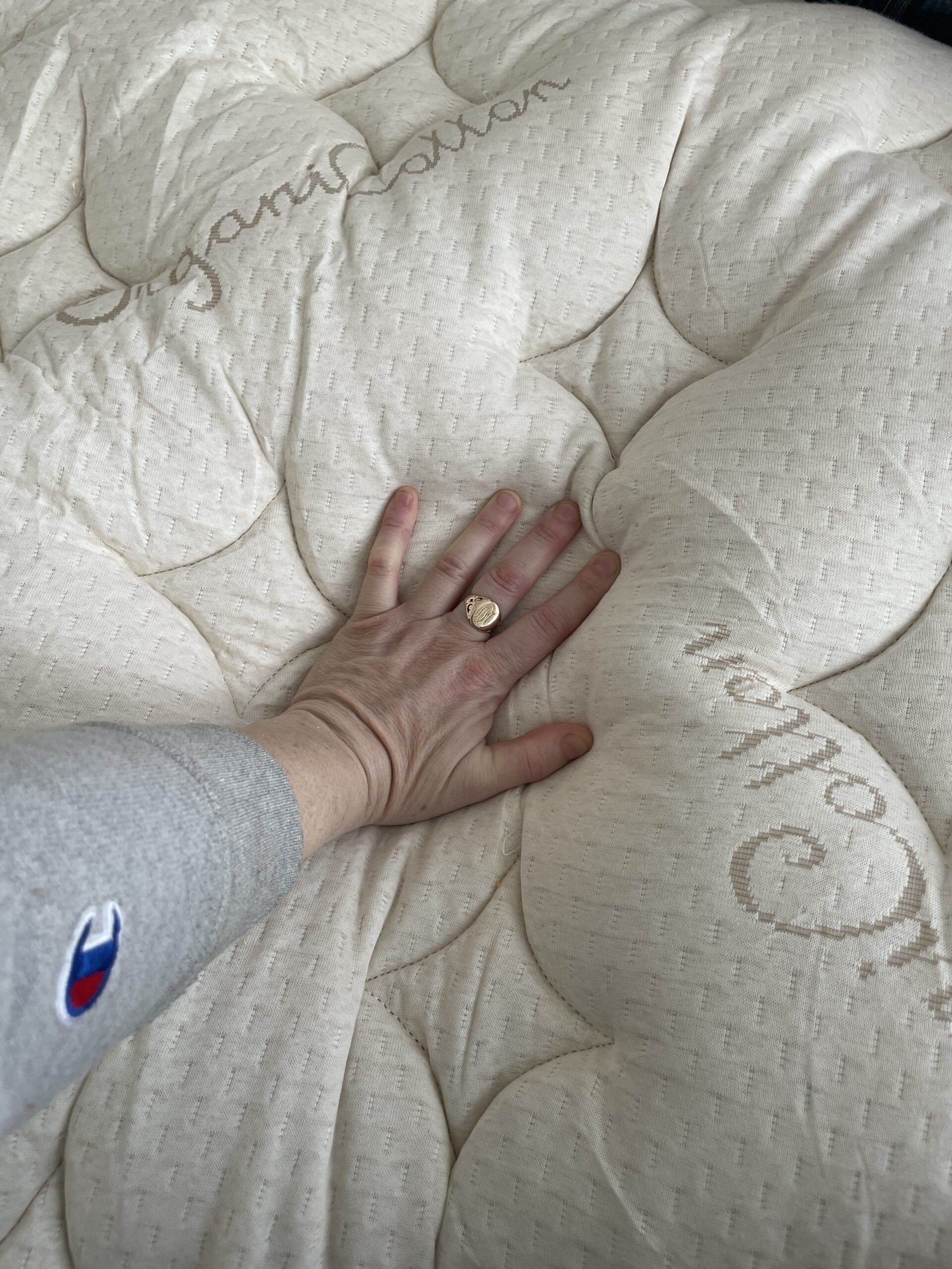 A hand pressing down on a textured, cushioned mattress. The mattress has stitched patterns and the words "organic" visible. The person is wearing a ring and a gray sleeve with a blue logo.