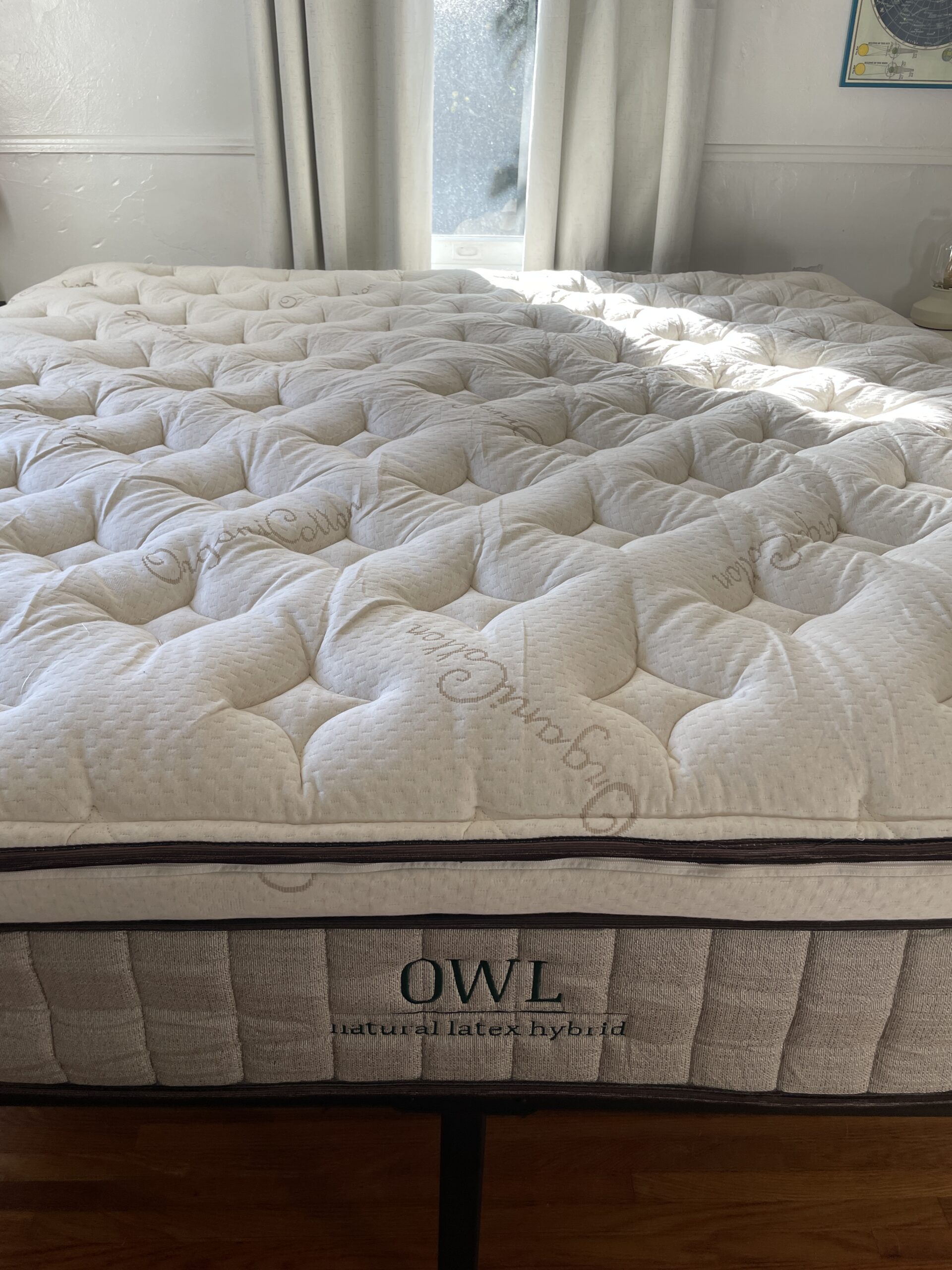 A neatly made bed with a quilted mattress labeled "OWL Natural Latex Hybrid" on a dark bed frame, positioned in a room with light-colored curtains and walls.
