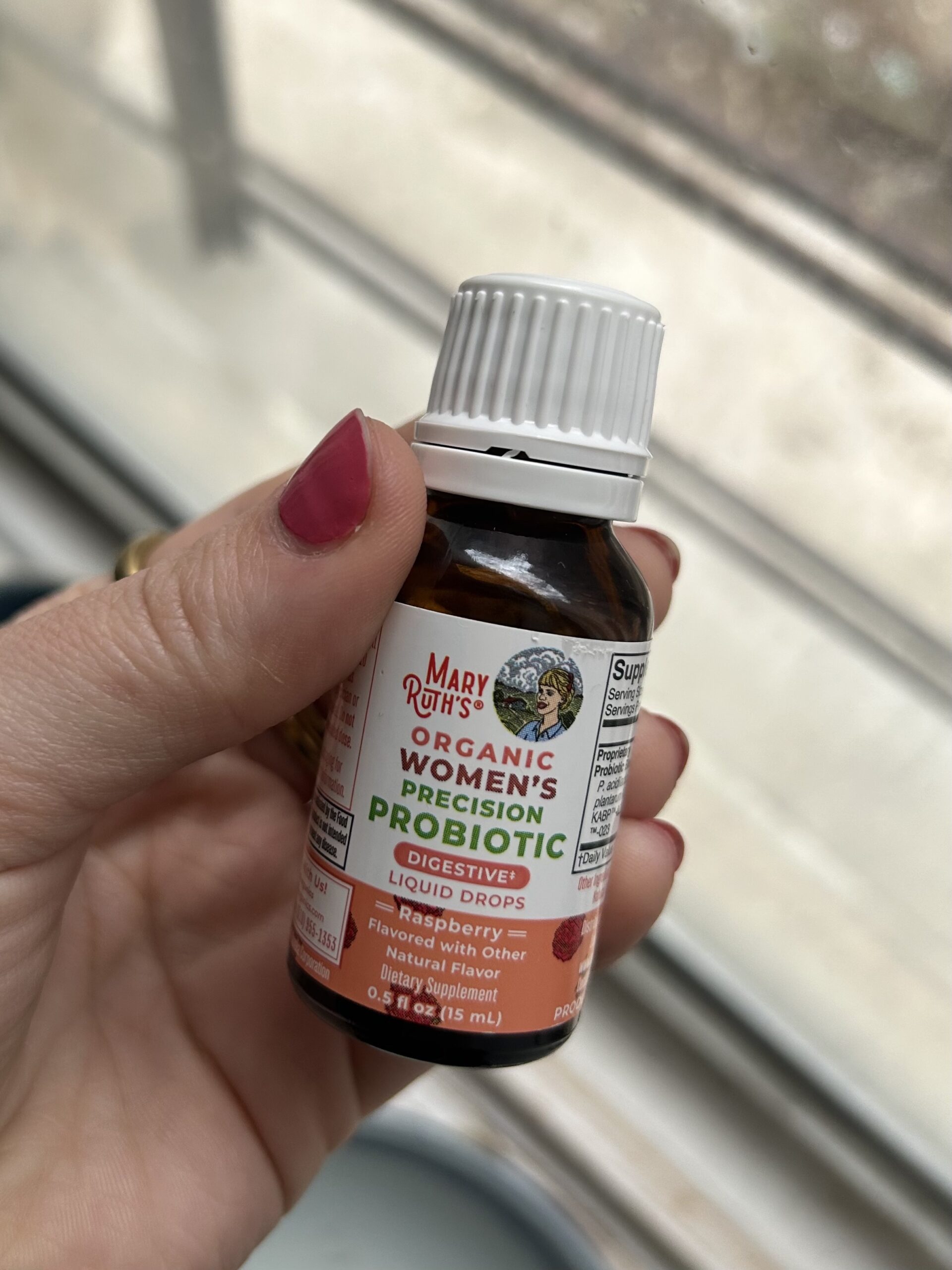 A hand holds a bottle of Mary Ruth’s Organic Women’s Precision Probiotic Digestive Liquid Drops, First Berry flavor, near a window.