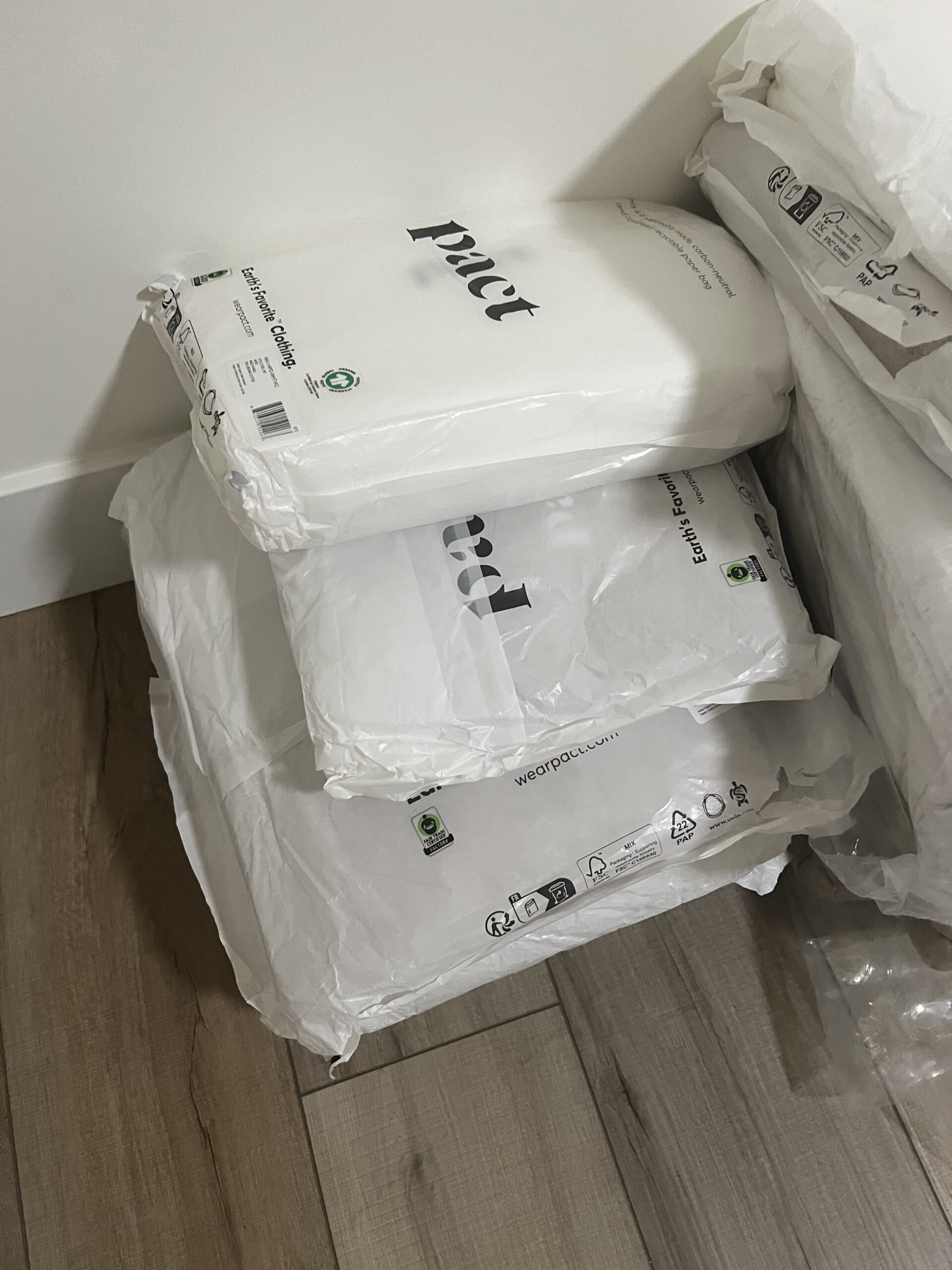 Stack of white packages with black text and recycling symbols, placed on a wooden floor next to a white wall.