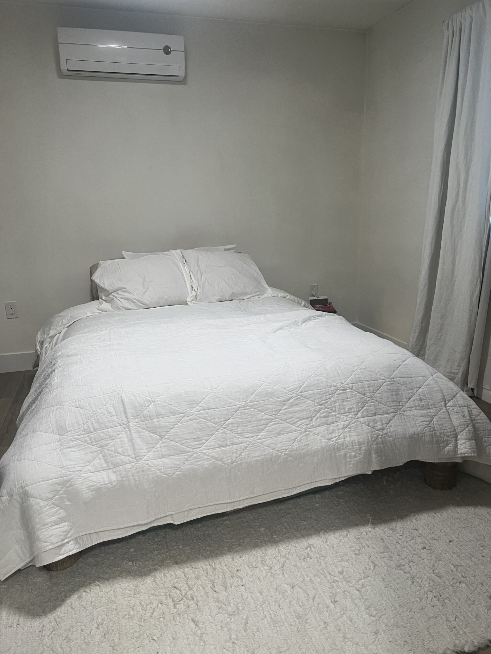 A neatly made bed with white bedding is in a room with light walls. There is an air conditioning unit above the bed and a window with white curtains to the right.
