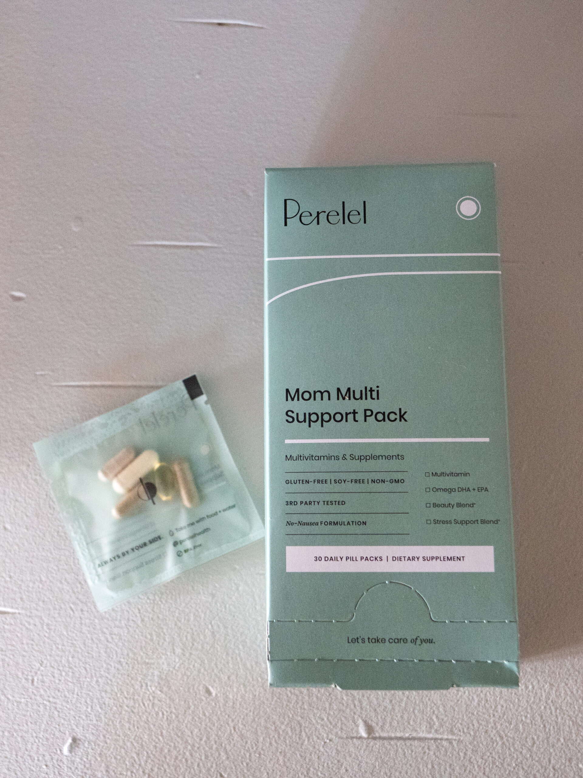 Box of "Mom Multi Support Pack" multivitamins with a small packet containing assorted pills, on a light surface.