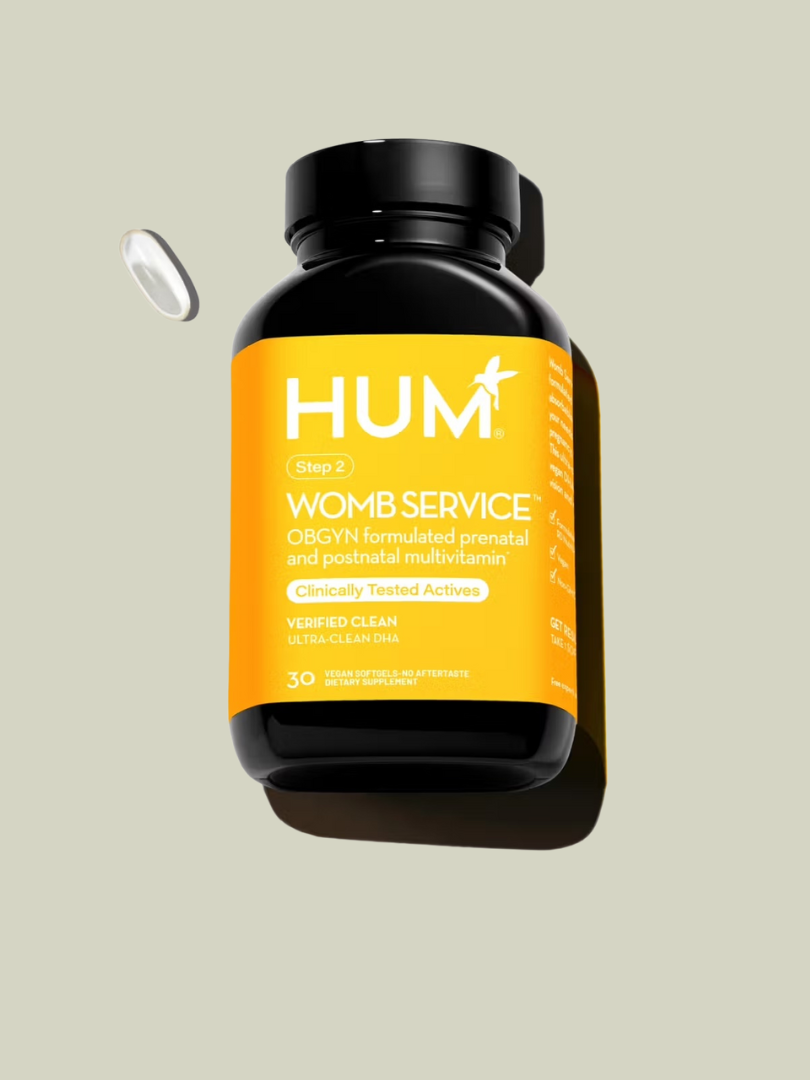 A bottle of HUM "Womb Service" prenatal and postnatal multivitamins with an orange label, floating against a light background. One capsule is shown beside the bottle.