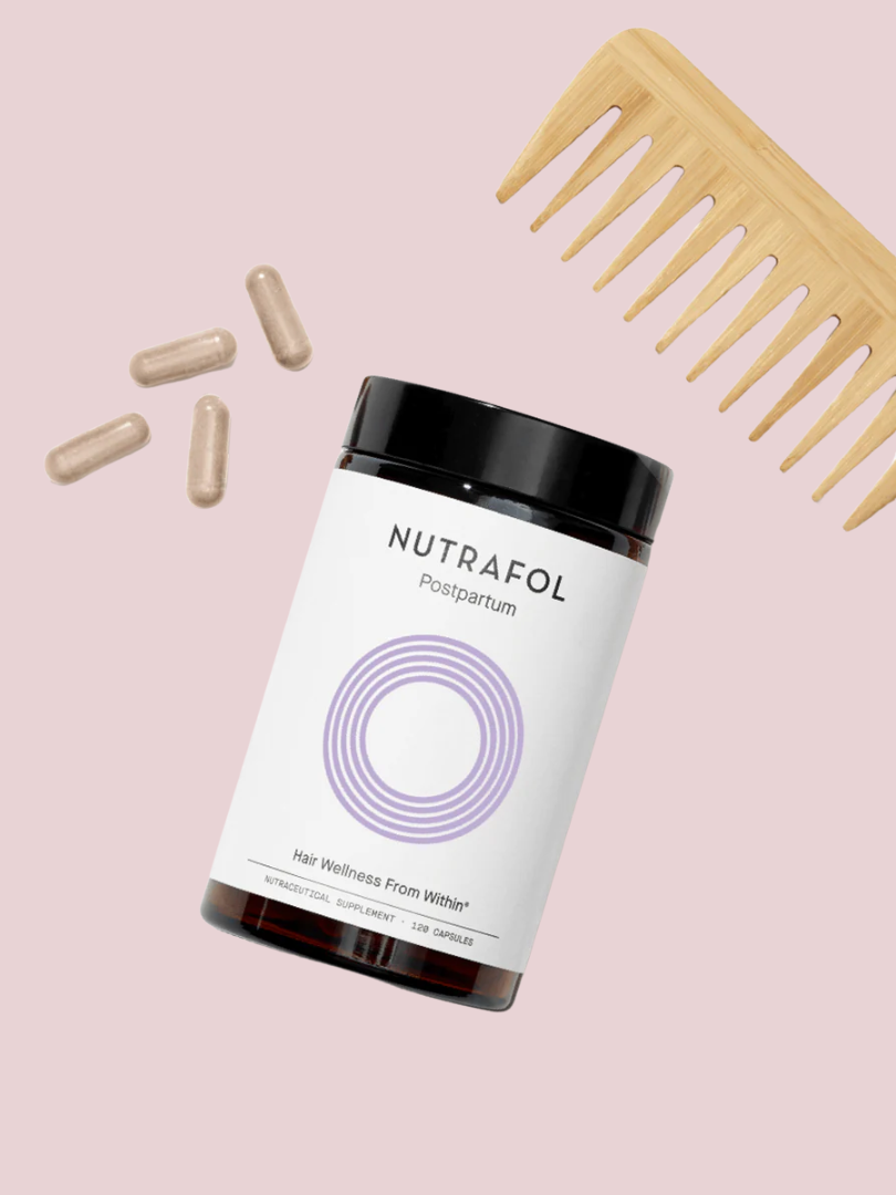 Nutrafol Postpartum hair supplement bottle, four capsules, and a wooden comb on a light pink background.