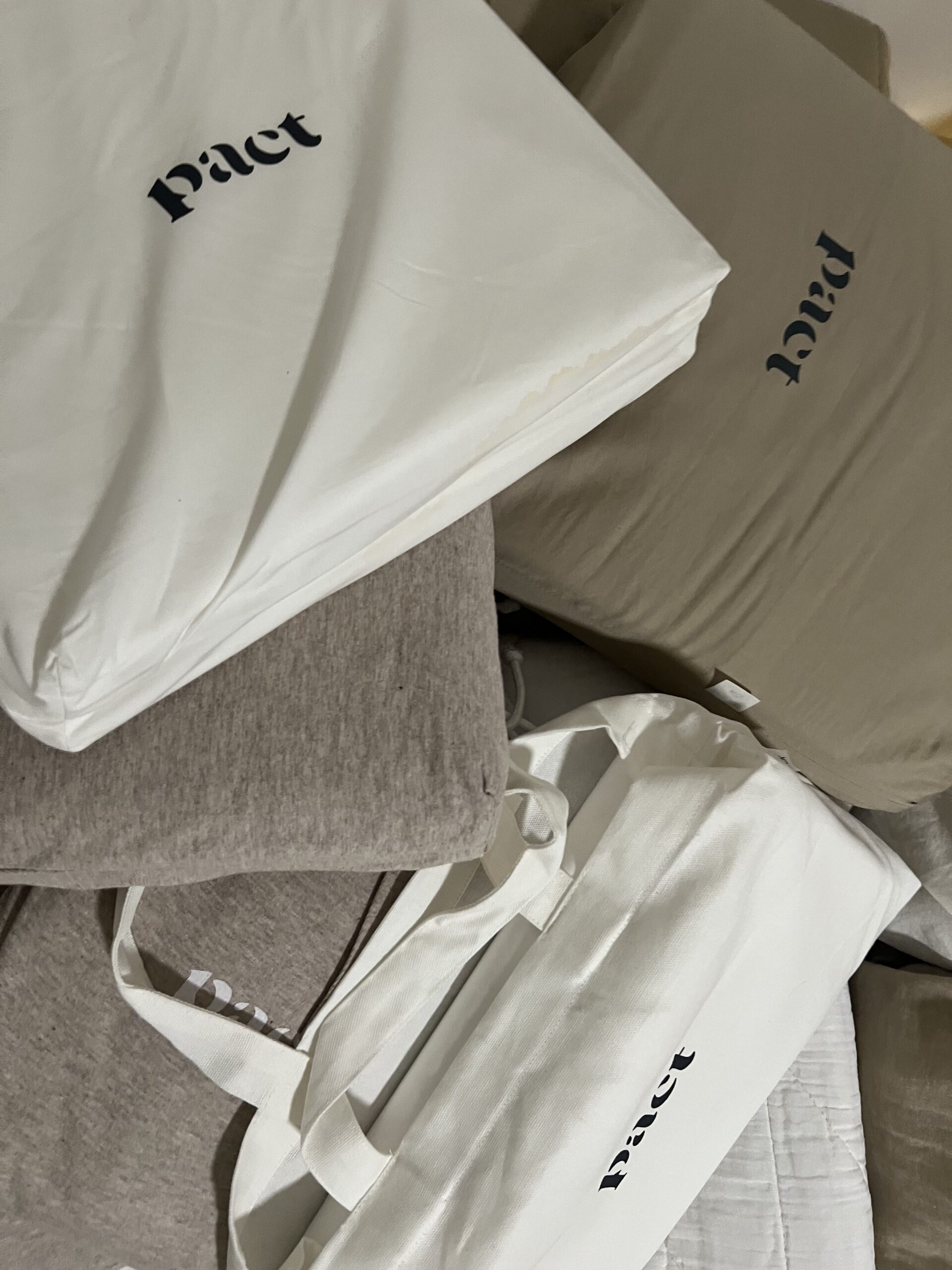 A pile of bags and pillows, some labeled "pact," in various shades of white, gray, and khaki green.