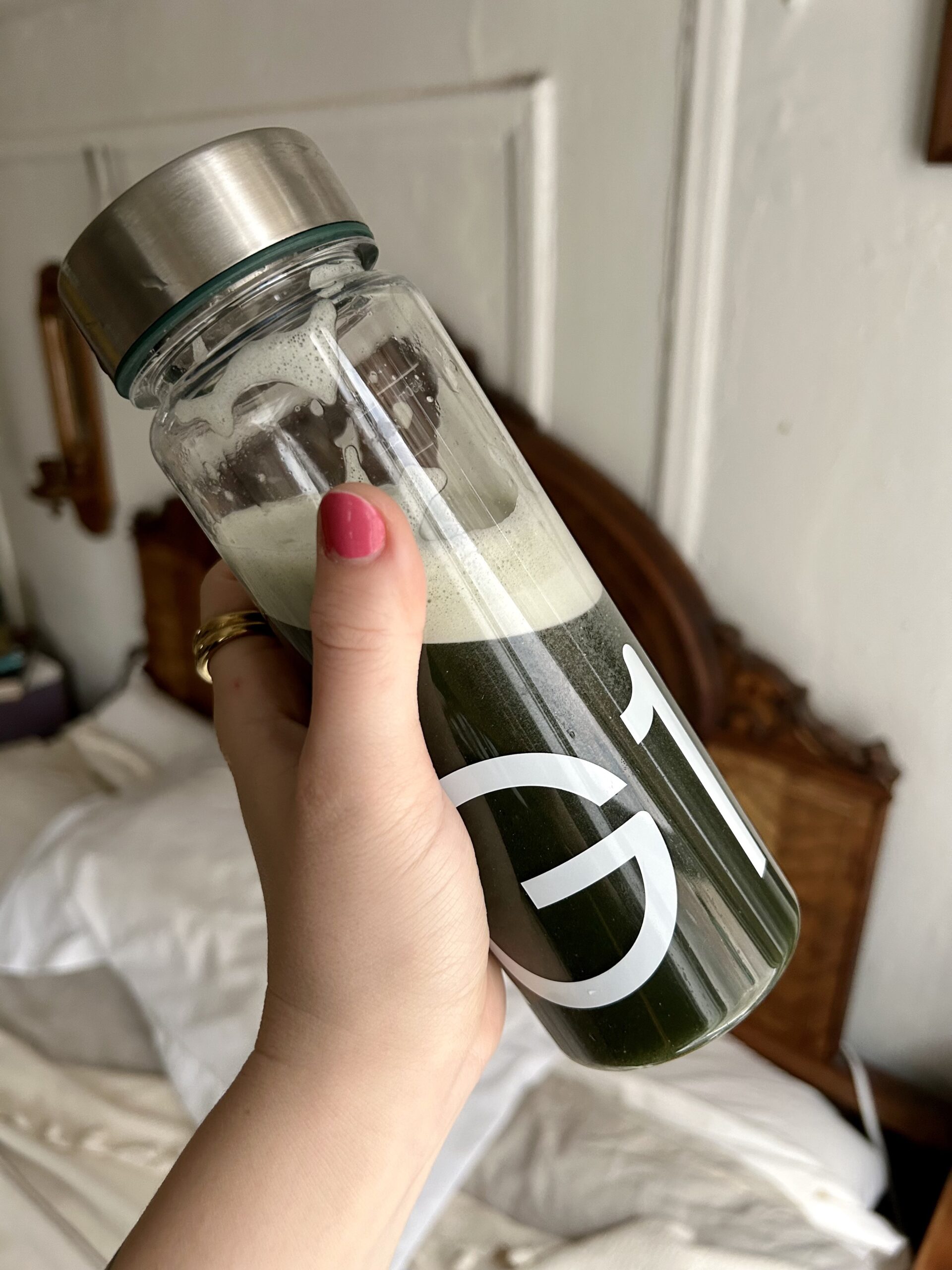 A hand with pink nail polish holds a glass bottle of green juice in front of an unmade bed and wooden headboard.