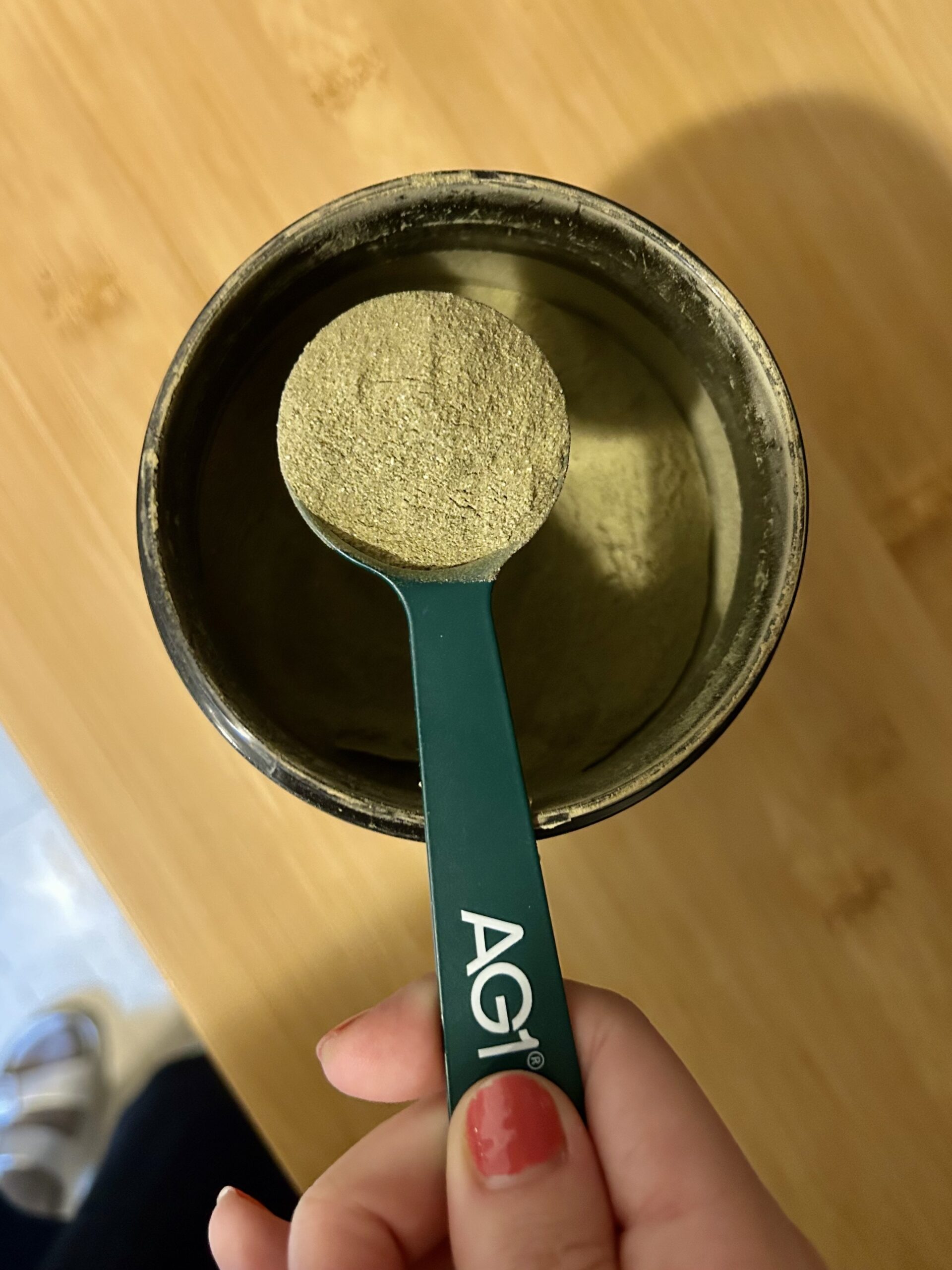 A hand holds a green spoon labeled "AG1" filled with greenish powder above an open container of the same powder on a wooden surface.