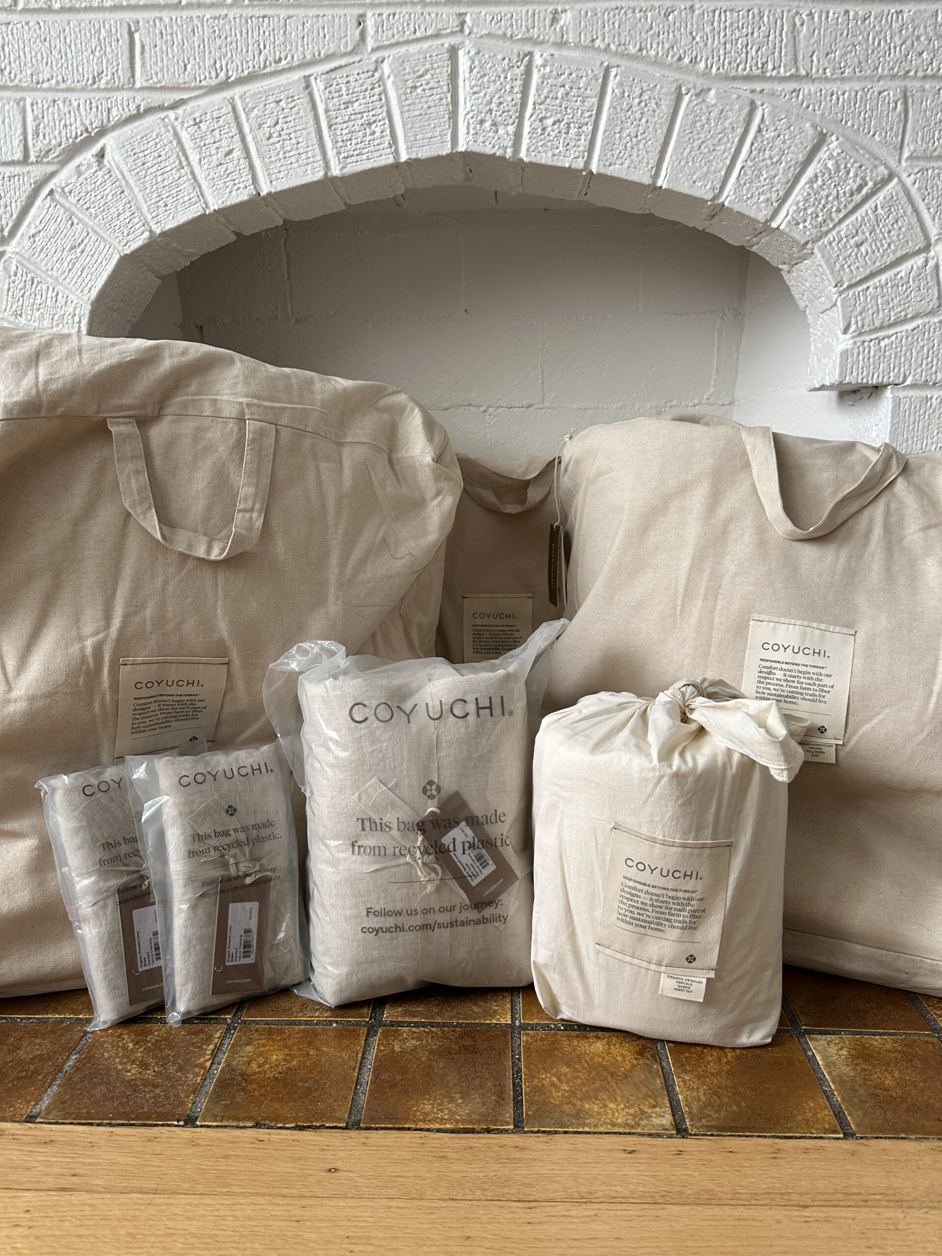 Assorted Coyuchi home textiles, including bags and packaged linens, displayed in front of a white brick fireplace.