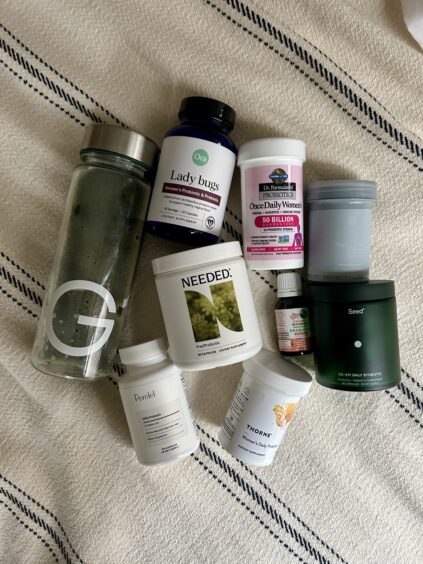 A glass water bottle and various supplement containers, including probiotics, vitamins, and powders, arranged on a striped fabric background.