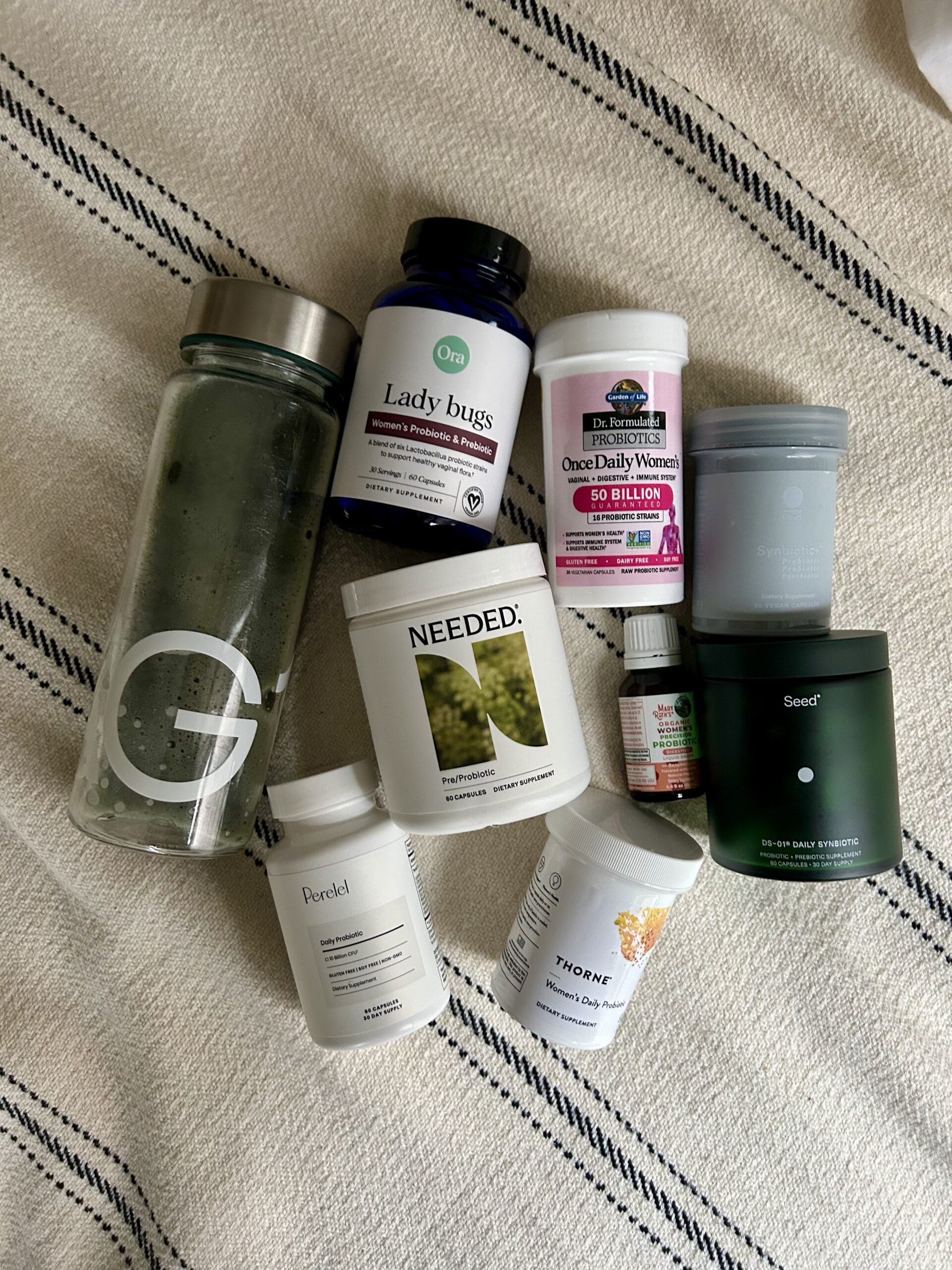A glass water bottle and various supplement containers, including probiotics, vitamins, and powders, arranged on a striped fabric background.