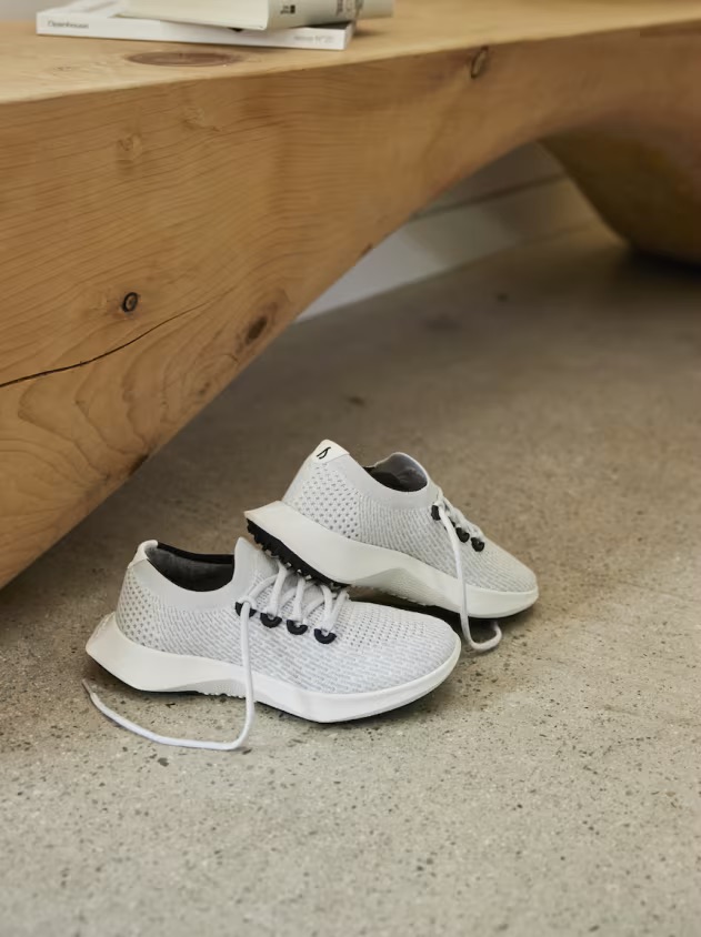 A pair of white athletic shoes with white laces on a concrete floor next to a wooden bench.