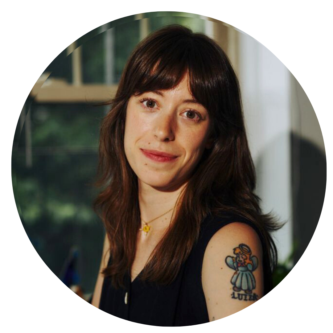 A woman with brown hair and bangs sits indoors, wearing a sleeveless top and a necklace, showing a tattoo of a cartoon figure labeled “Lulu” on her upper arm.