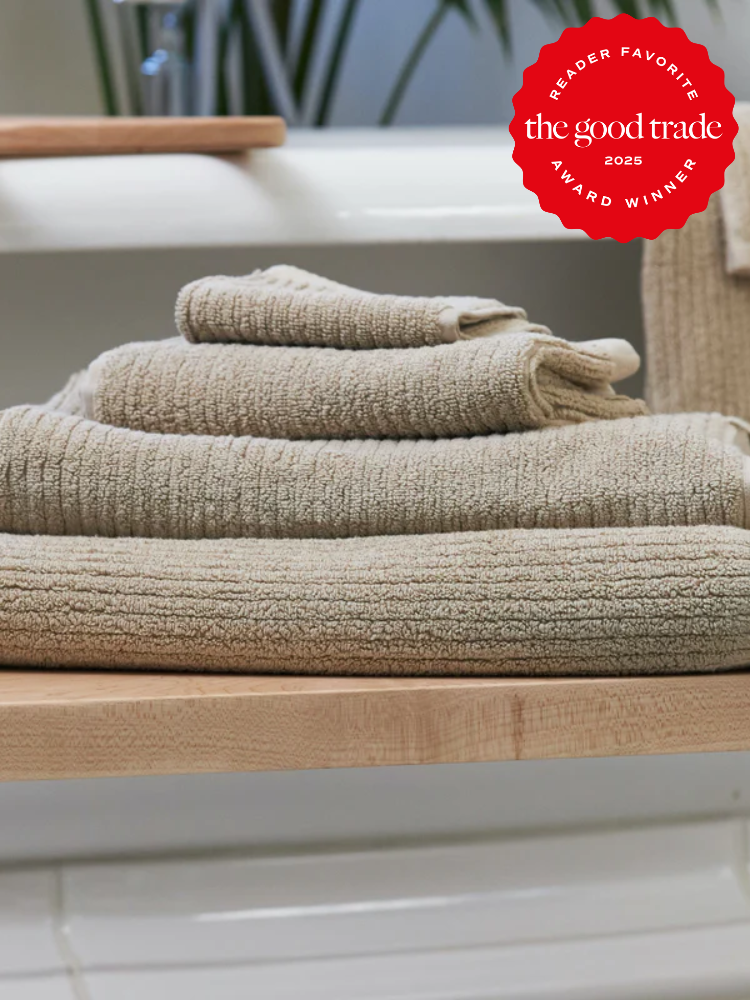 A stack of beige ribbed organic towels folded on a wooden shelf, with a red "The Good Trade Reader Favorite Award Winner 2025" badge in the corner.