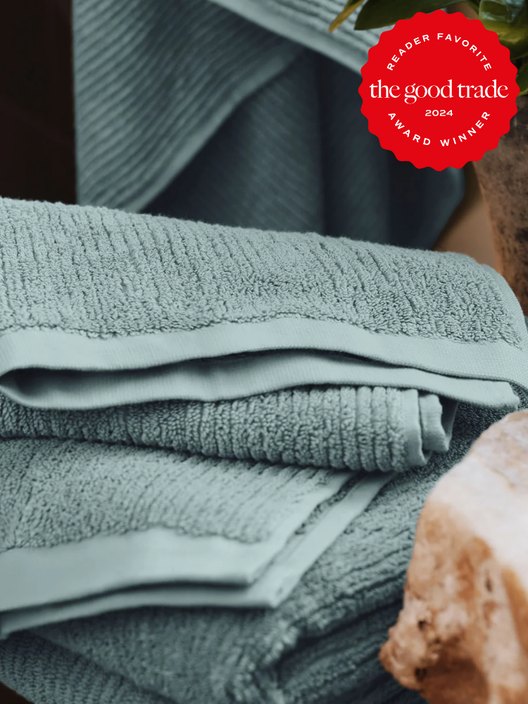 A stack of folded light blue organic towels sits on a surface, with a red award badge in the corner reading “The Good Trade Reader Favorite Award Winner 2024.”.