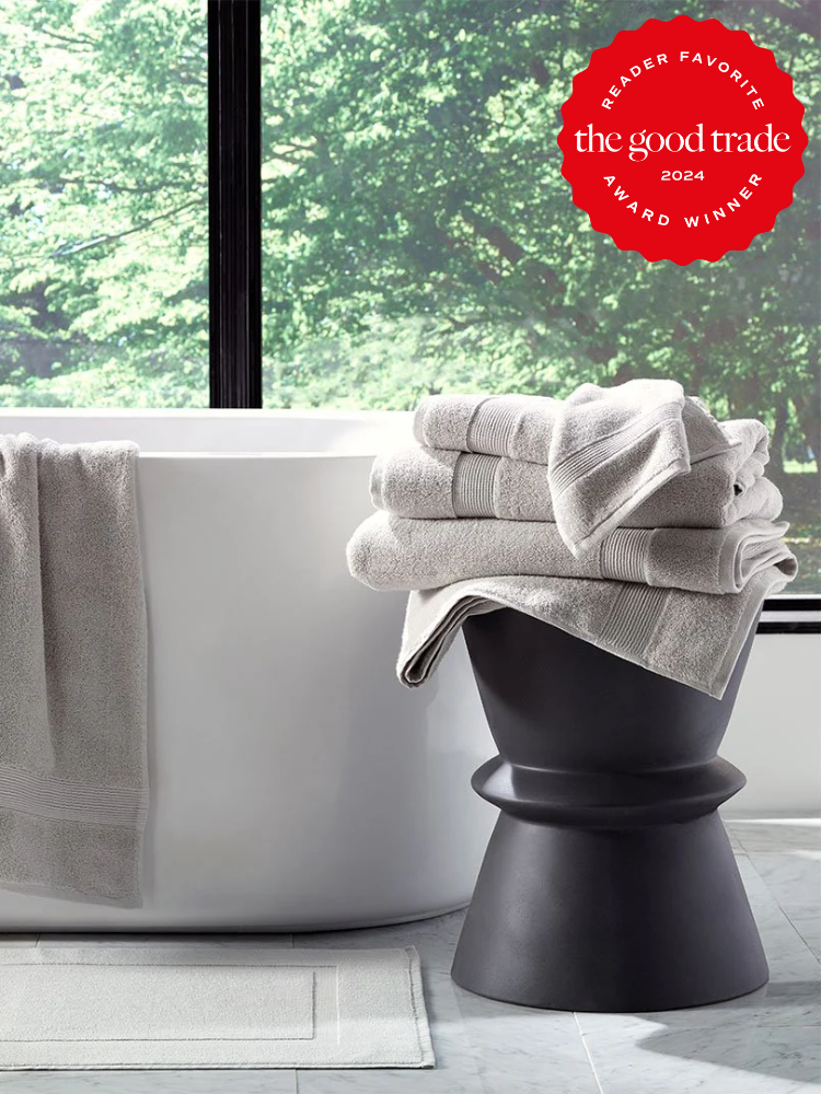 A white freestanding bathtub sits beside a black stool stacked with folded organic towels, with a large window revealing lush greenery outside.