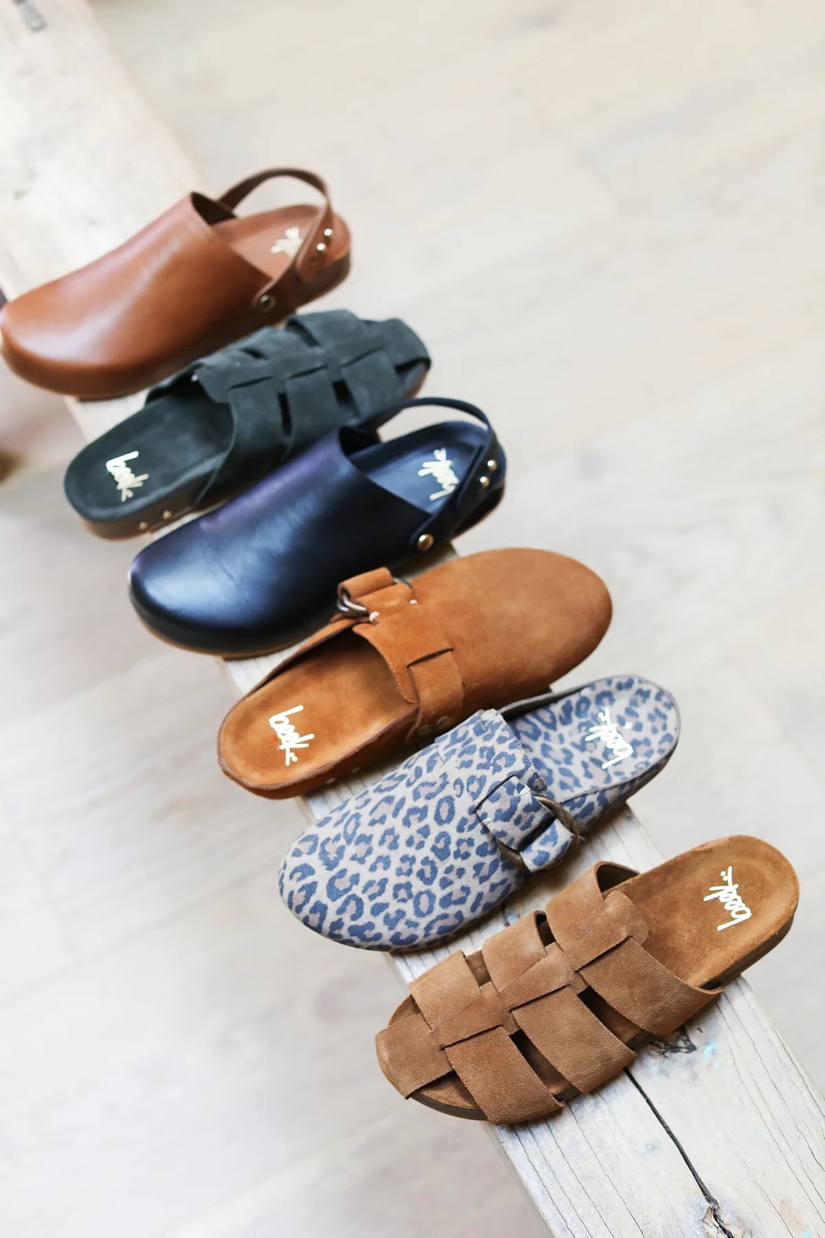 Five pairs of slip-on sandals in various colors and patterns, including brown, black, navy, suede, and leopard print, arranged on a wooden surface.