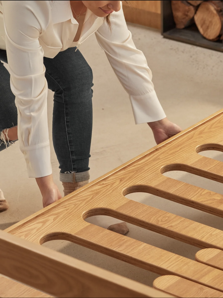 A person in a white shirt and jeans assembles a wooden bed frame, placing slats into position in a bright room with firewood in the background.