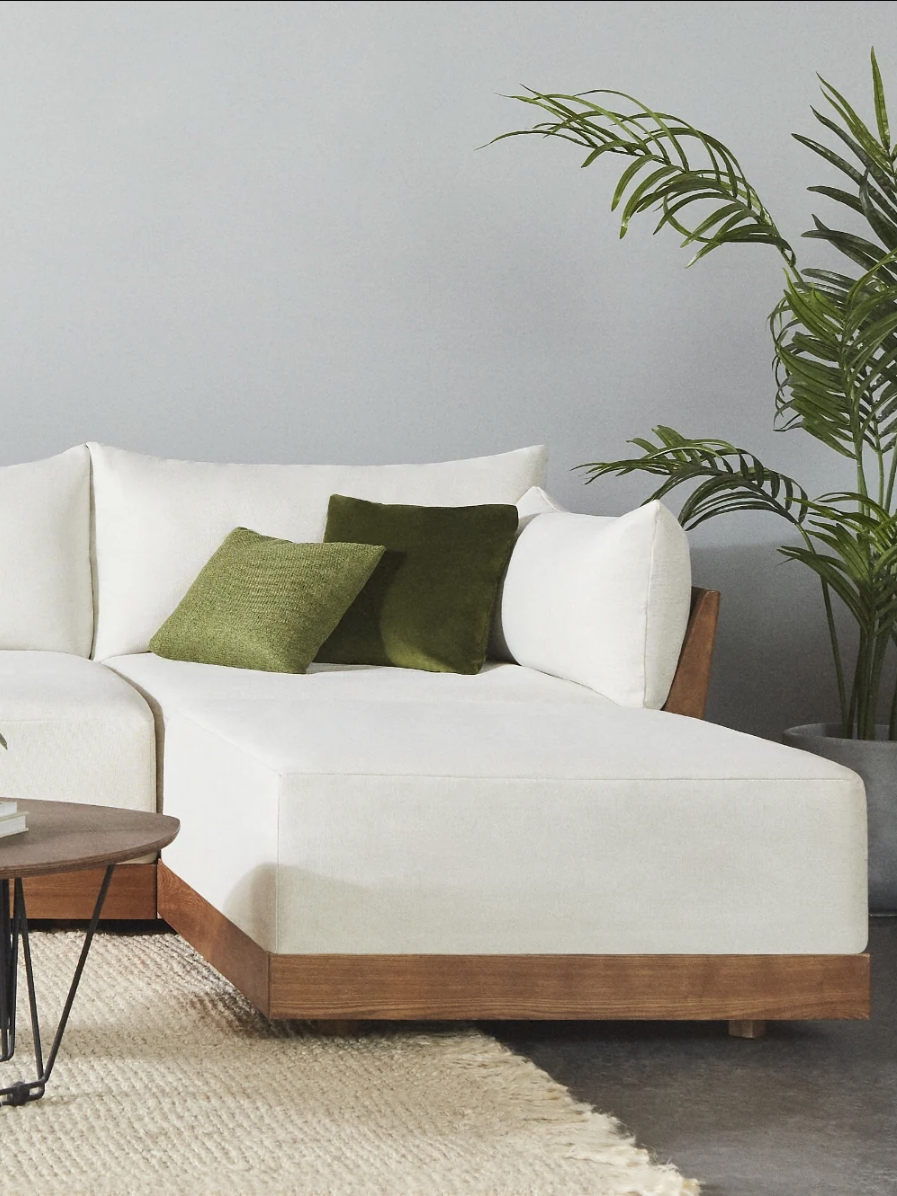White sectional sofa with two green cushions, wooden base, next to a large potted plant. A round table holds a small plant and book. Beige rug on the floor. Neutral wall in the background.