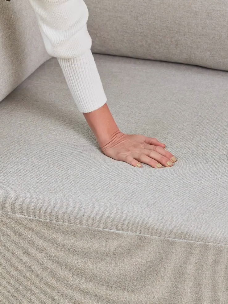 A hand presses down on a light gray couch cushion, showing the fabric and texture.