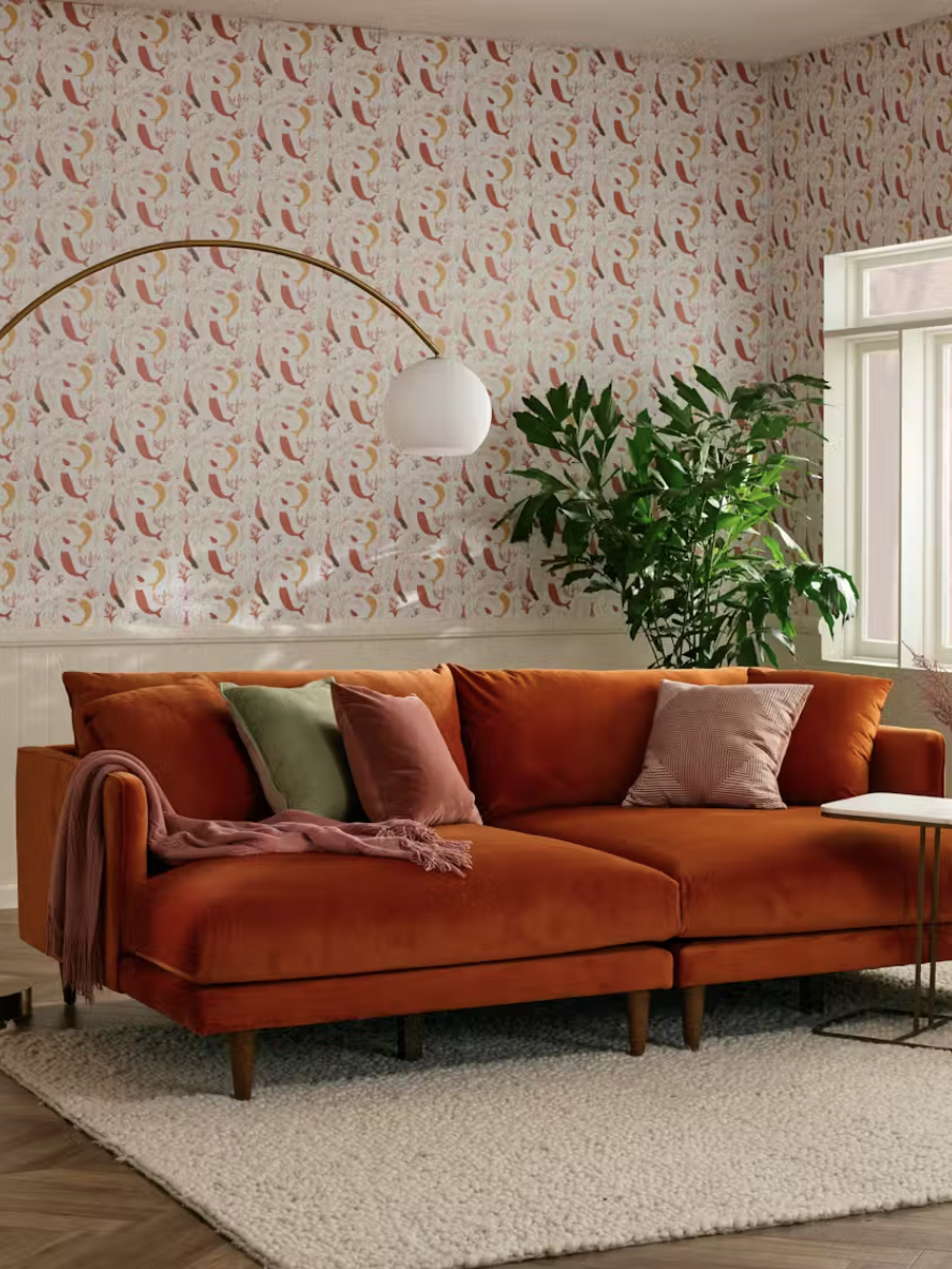 Living room with a rust-colored sofa, pillows, a white floor lamp, a potted plant, and patterned wallpaper. A small side table and large window are also visible.