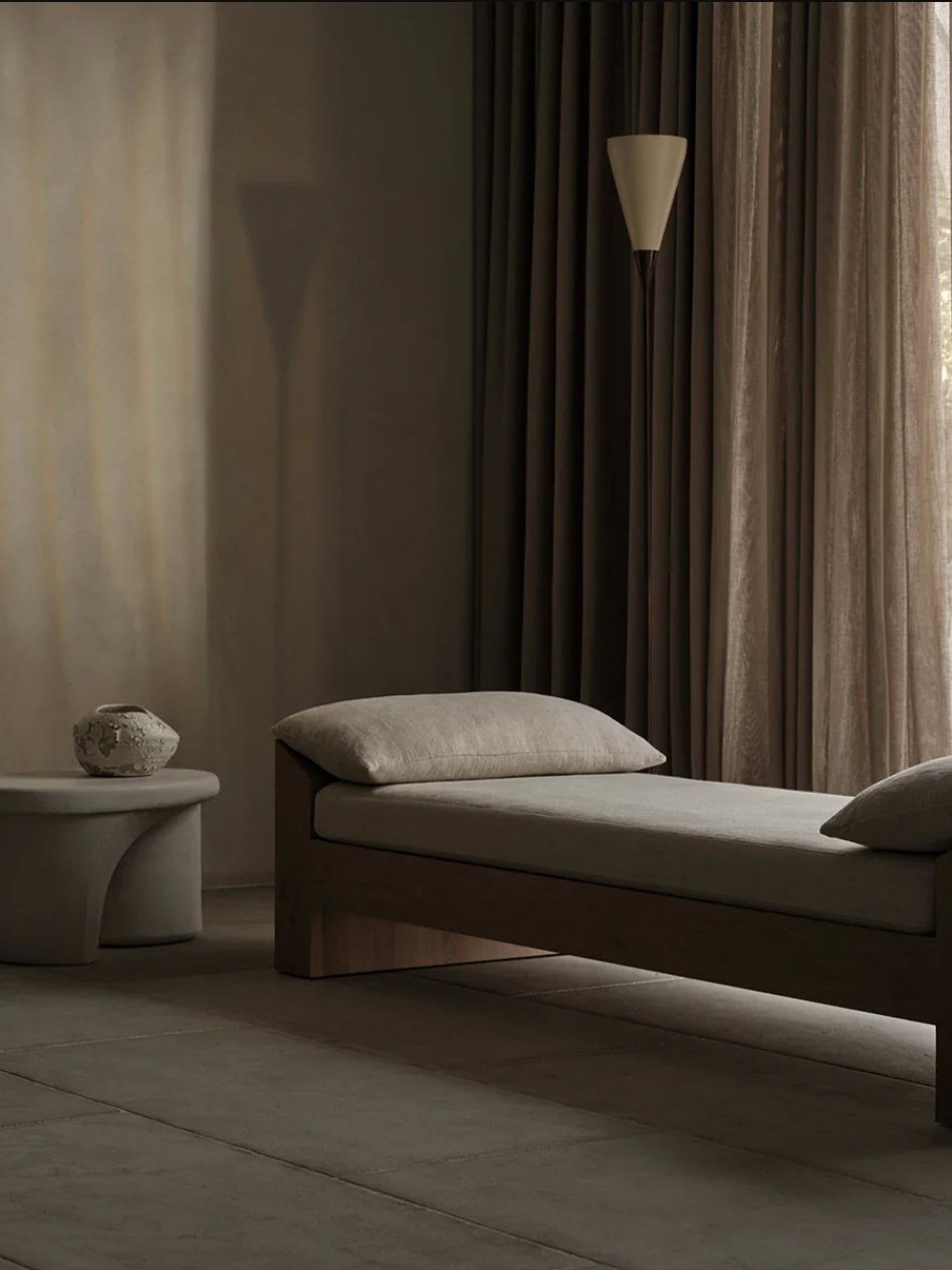 A minimalist room with a beige daybed, a small round side table holding a decorative stone object, and tall drapes in the background.