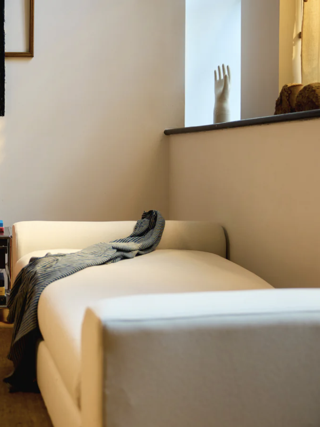 A cozy living room corner with a white sofa draped with a striped blanket, a small side table with books and decor, a window ledge with a hand sculpture, and warm lighting.