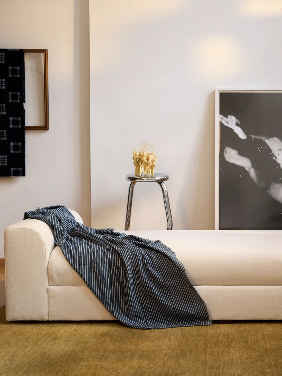 Minimalist room with a white daybed, striped blanket, stool with decorative item, framed photographs on walls, and green carpet.