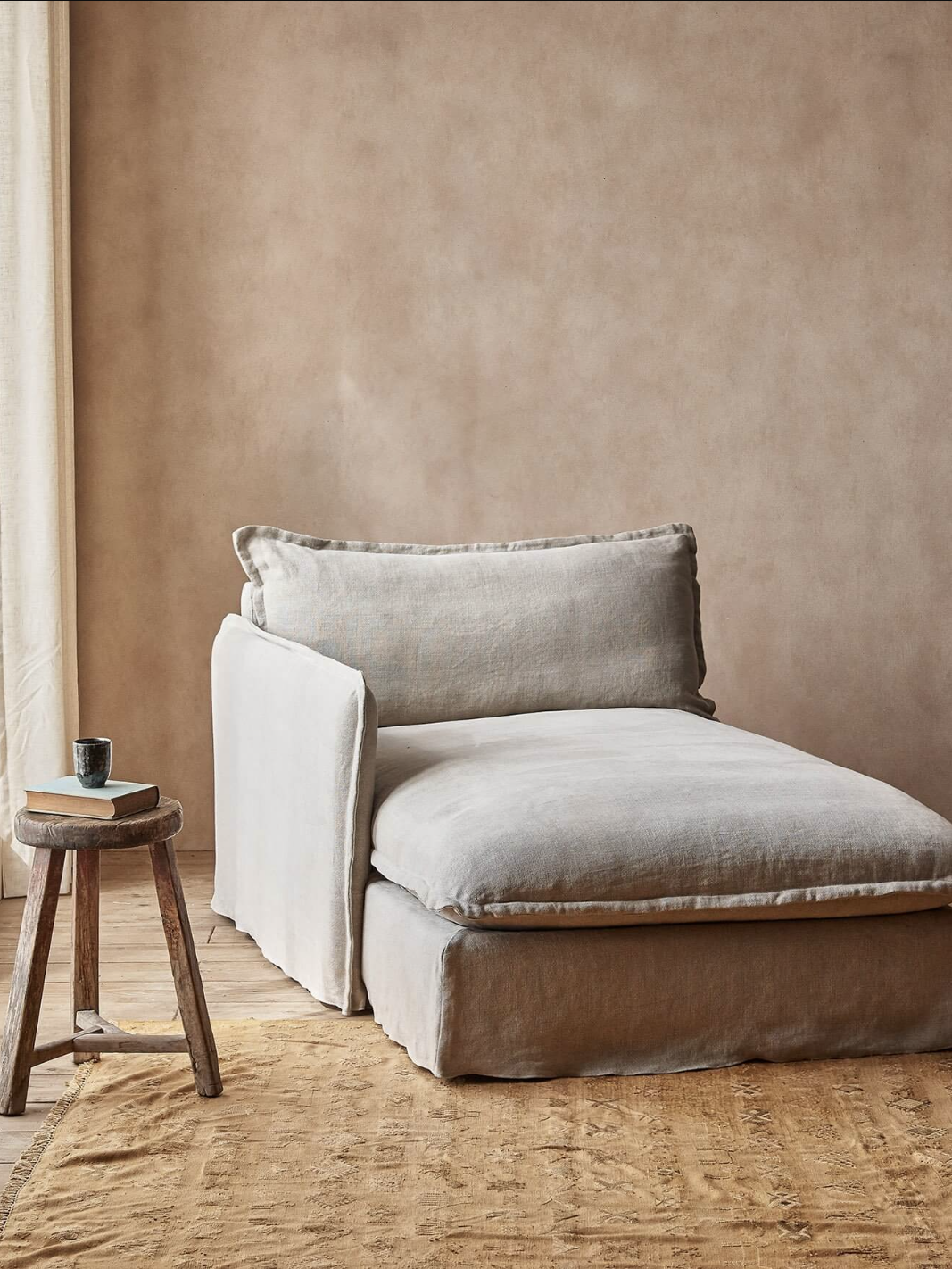 A beige chaise lounge with a cushioned backrest is placed on a tan rug next to a small wooden stool with a coffee cup. Light filters in from a window, illuminating the setup.