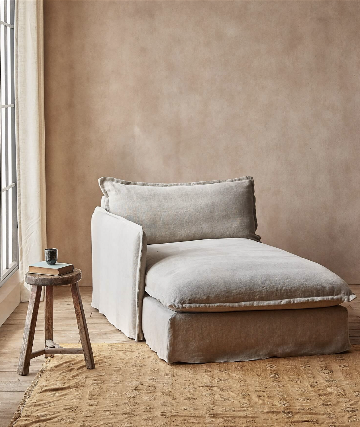 A beige chaise lounge with a cushioned backrest is placed on a tan rug next to a small wooden stool with a coffee cup. Light filters in from a window, illuminating the setup.