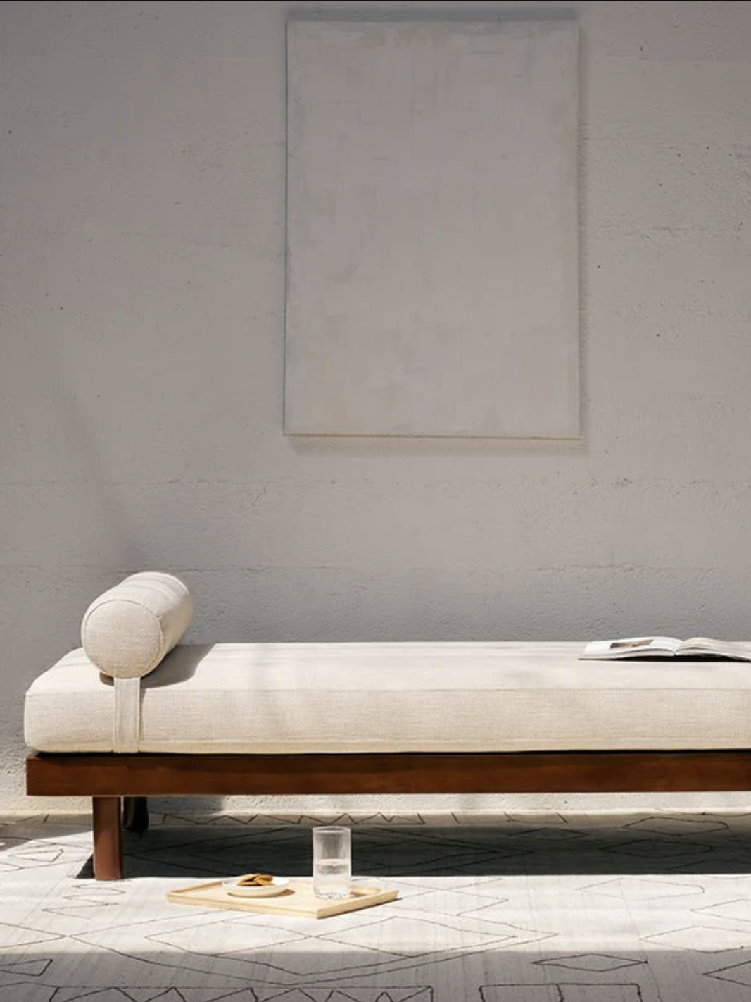 Minimalist room with a wooden daybed featuring a cylindrical pillow, open book, and a tray with a glass of water and a snack. A blank canvas hangs on the wall behind.