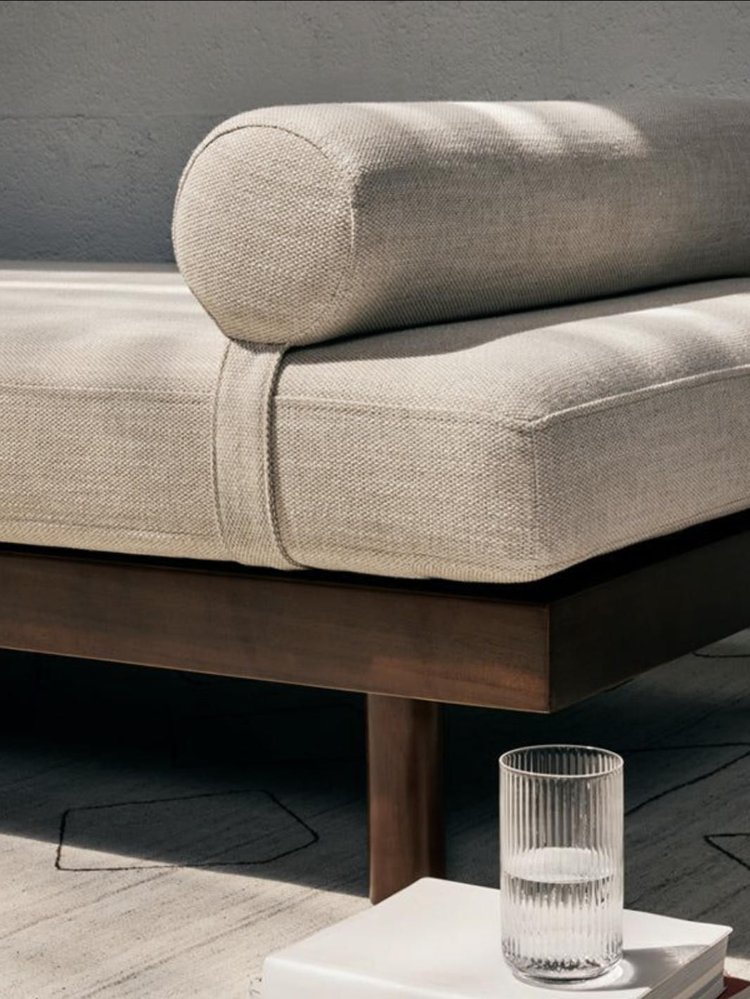 A beige upholstered daybed with a cylindrical pillow on a wooden frame. A glass of water and a stack of books sit on the ground beside it.