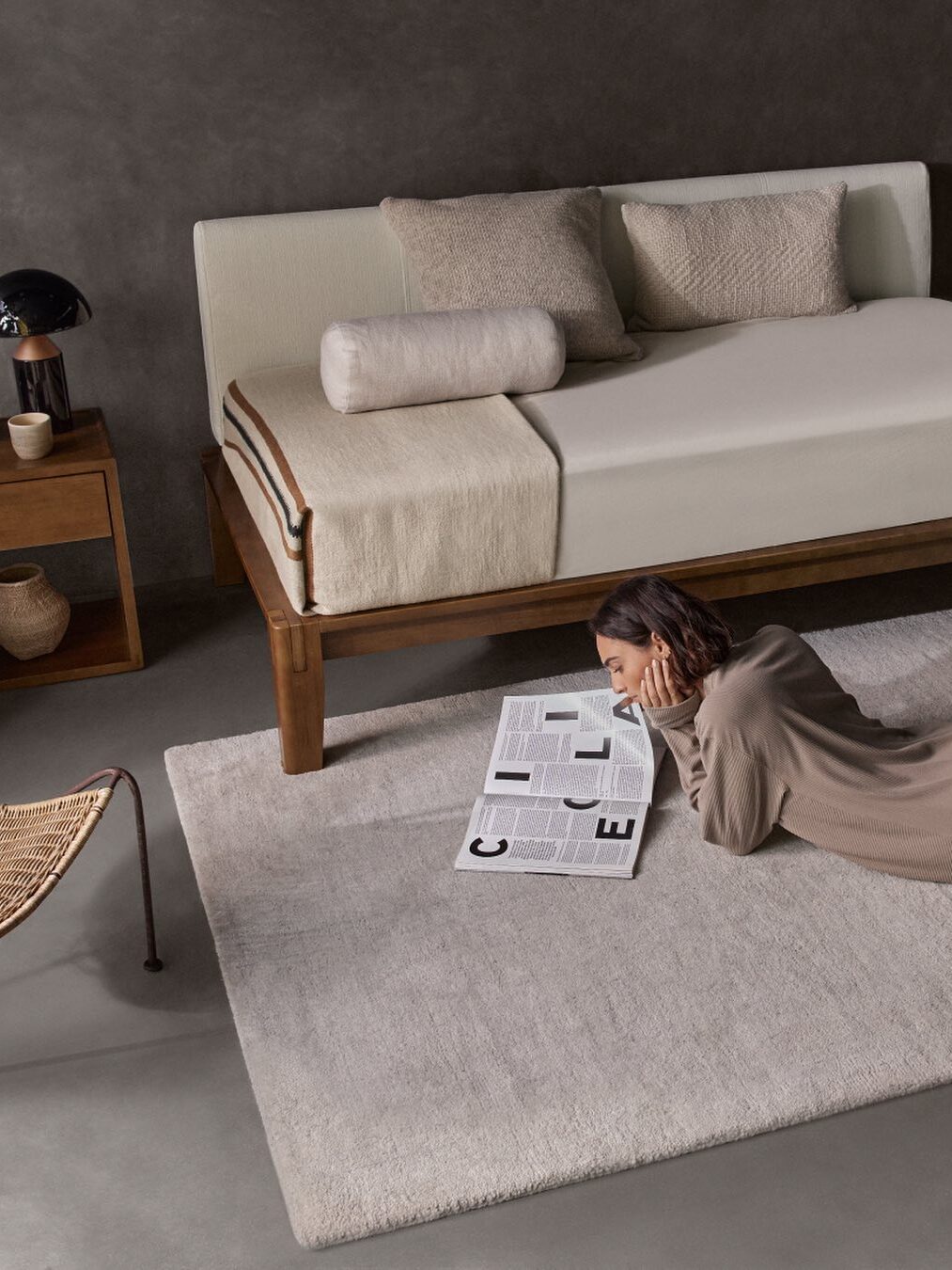Woman reading a newspaper on a beige rug beside a bed with a wooden frame, neutral-toned bedding, and a side table with a lamp and cups.
