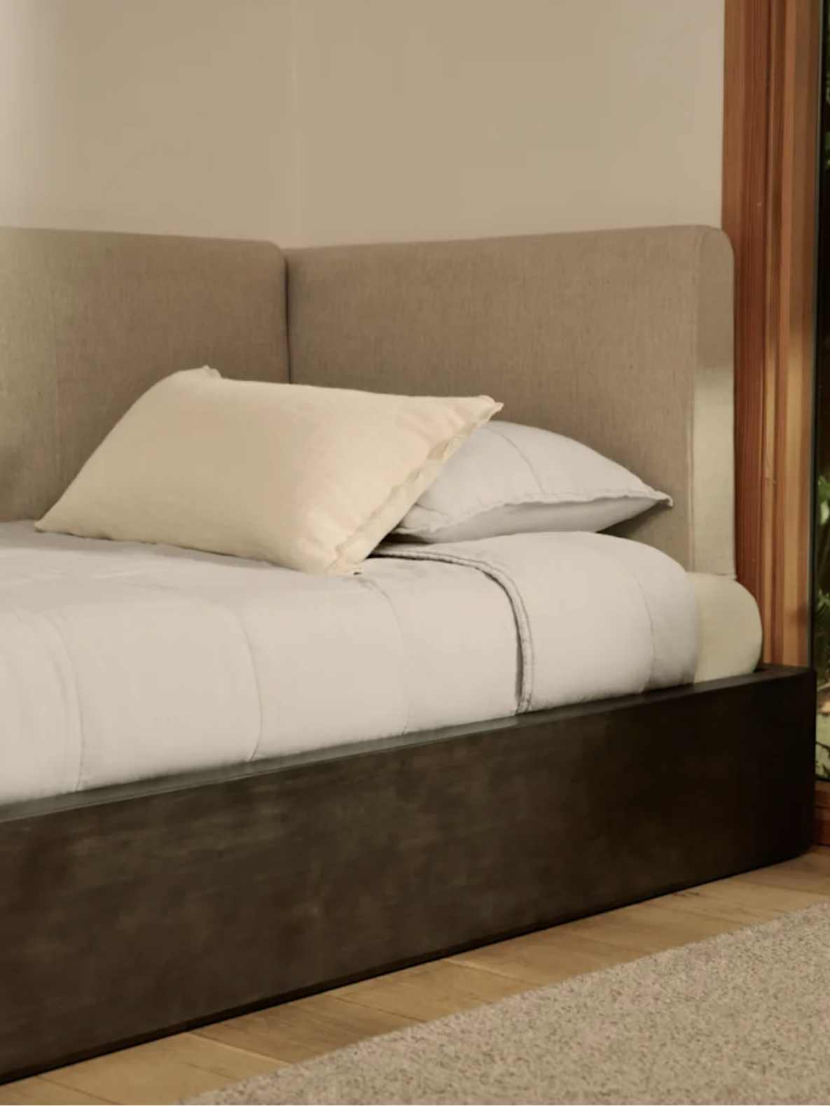 A neatly made bed with a beige headboard is positioned next to a window showing green plants outside.