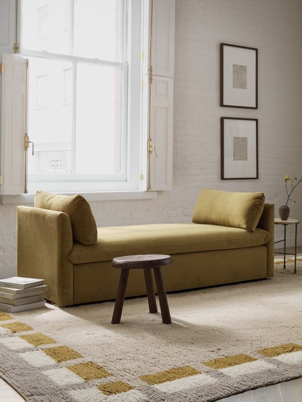 A minimalist living room with a mustard velvet daybed, a small wooden stool, two framed artworks on the wall, and a large window. A neutral-toned rug covers the floor.