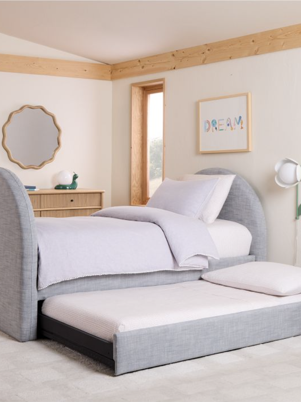 A bedroom with a gray upholstered bed featuring a pull-out trundle. There is a dresser with a round mirror and a plant lamp. A "DREAM" sign hangs on the wall.