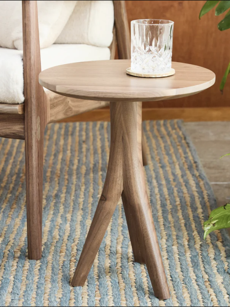 A small wooden side table with a round top and three legs, placed on a striped rug. A glass with a textured surface sits on the table.