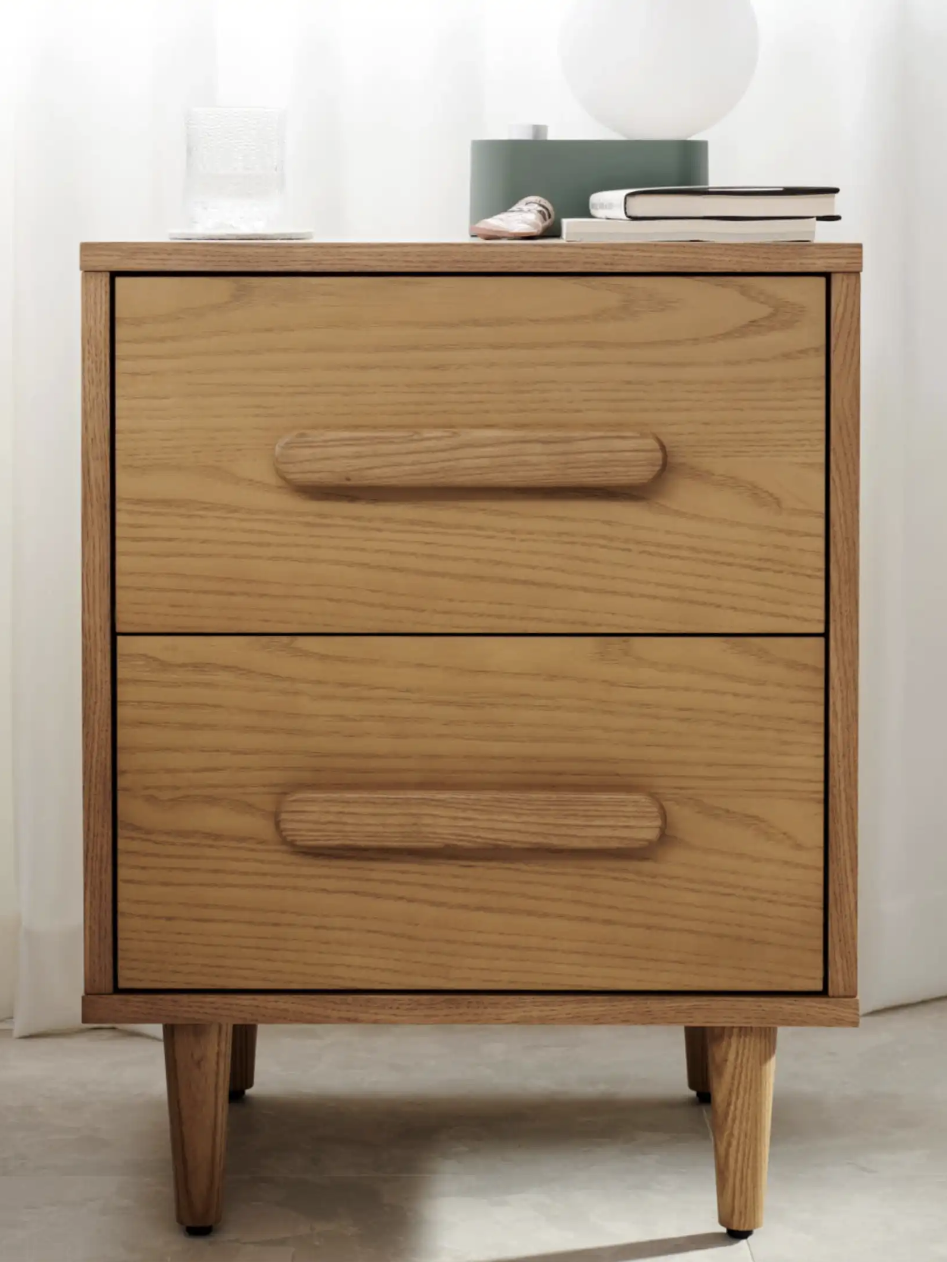 Wooden nightstand with two drawers, featuring large handles. A glass, book, and lamp are on top. The setting includes a white curtain and part of a bed on the left.