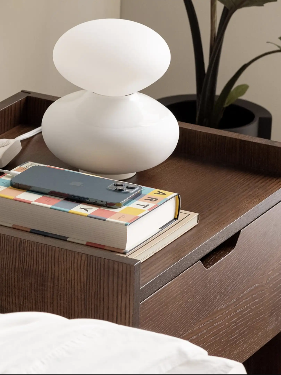 A wooden nightstand with a white lamp, a smartphone on a colorful book, and a power strip.