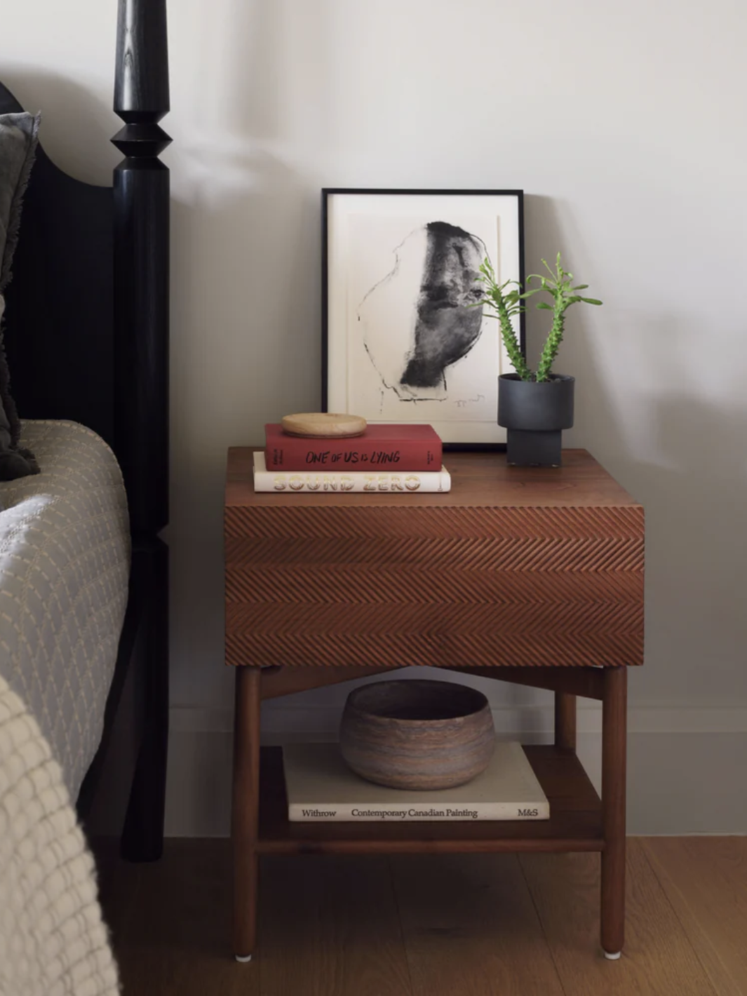 A bedside table with a potted plant, framed abstract artwork, and books, against a light wall. A bed with a patterned pillow and gray bedding is partially visible beside it.