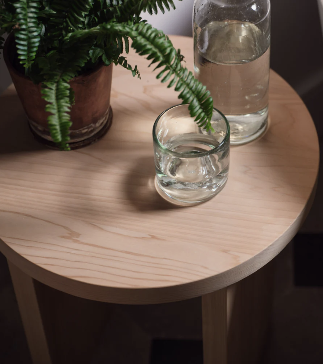 A round wooden table with a potted fern, a glass of water, and a glass bottle, partially filled, sitting on its surface.