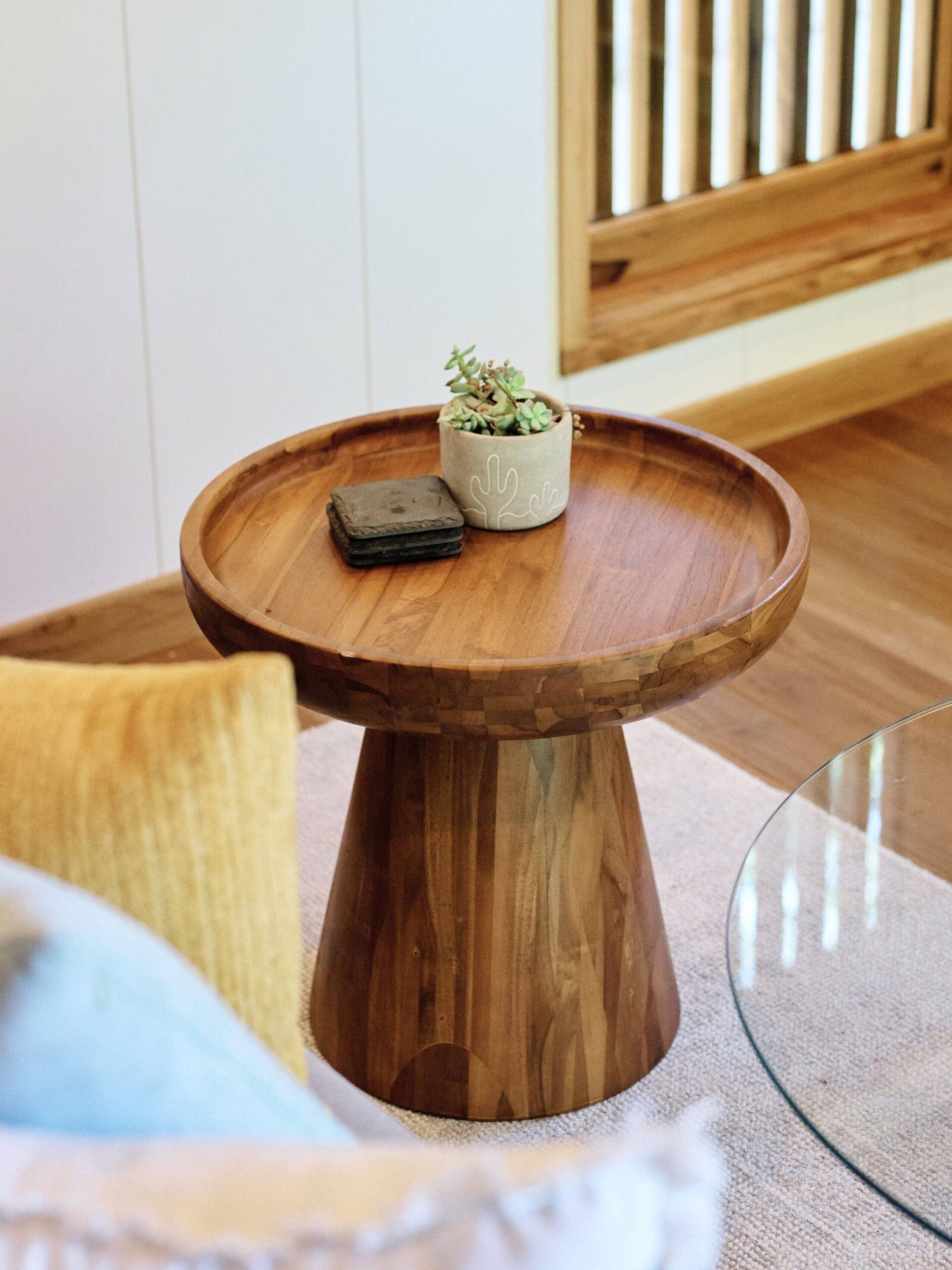 Round wooden side table with a small potted plant and a wallet on top, placed near a glass coffee table and cushions in a living room setting with a wooden floor.