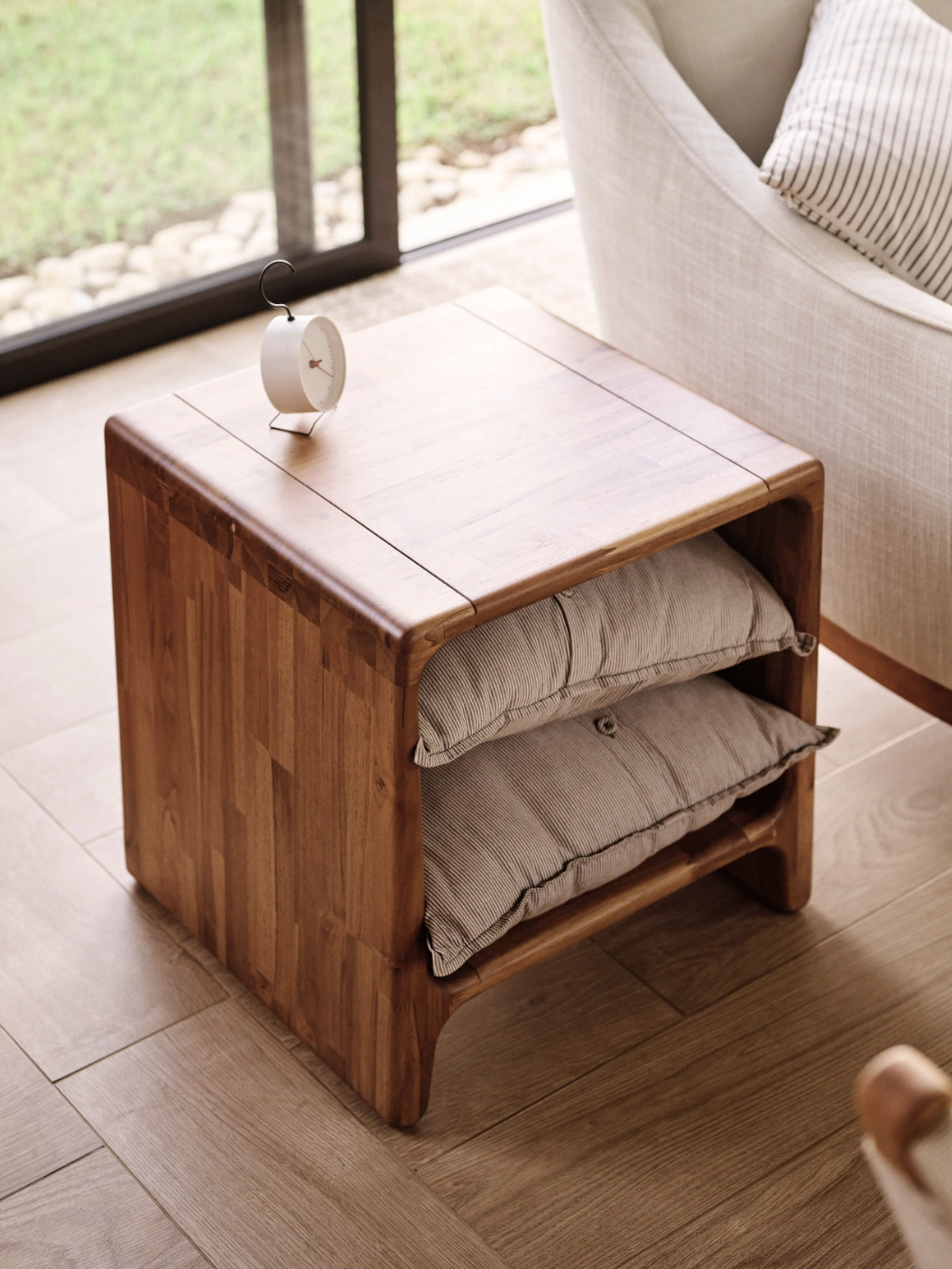 Wooden side table with two gray cushions stored underneath, next to a beige armchair and a small clock on top. Positioned on wooden floor by a window with a garden view.